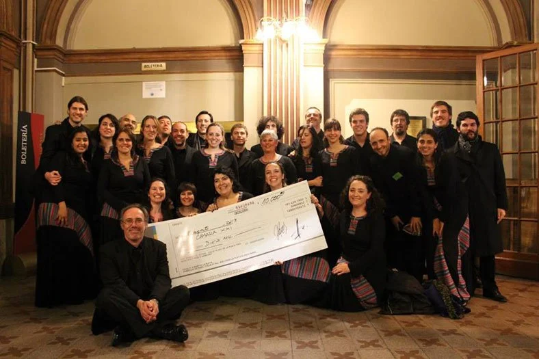 Grupo de personas sonriendo, algunas con vestimenta tradicional, sosteniendo un cheque grande en un salón con decoraciones clásicas.