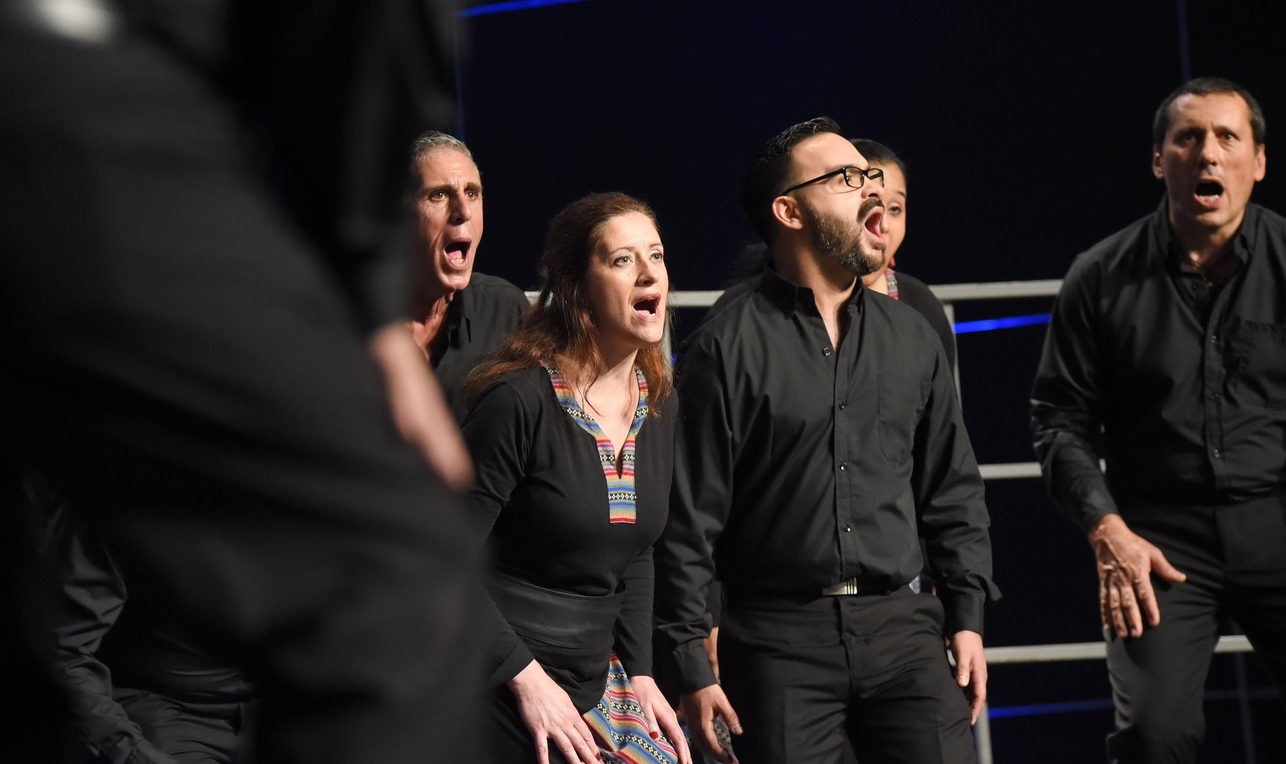Varias personas cantando o actuando en un escenario, con expresiones emocionales y vestidas con camisetas negras, en un ambiente de teatro o presentación.
