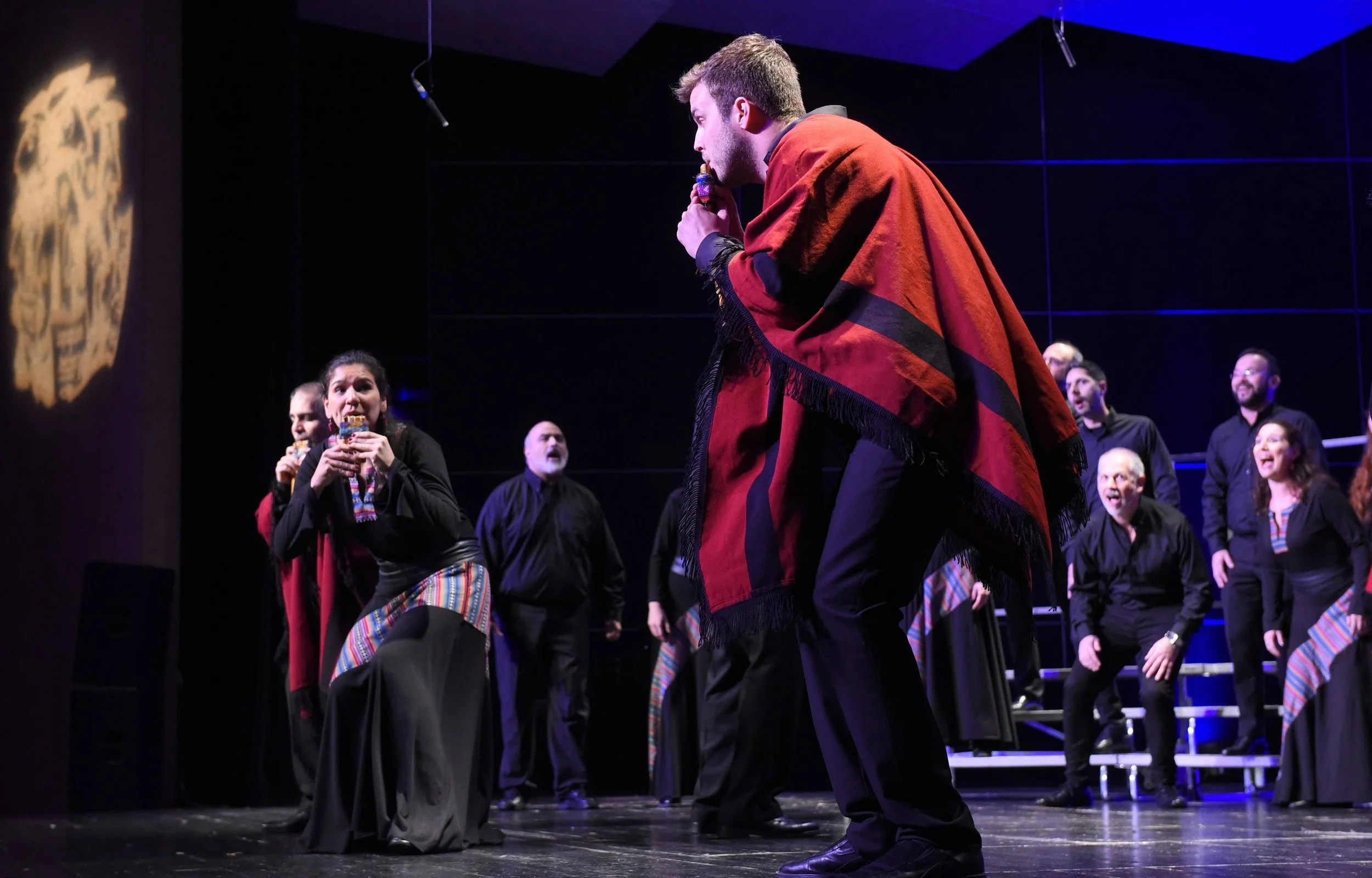 Un grupo de personas en un escenario, con vestimentas tradicionales, participando en una presentación o espectáculo, algunos sosteniendo objetos y otras cantando o dando instrucciones.