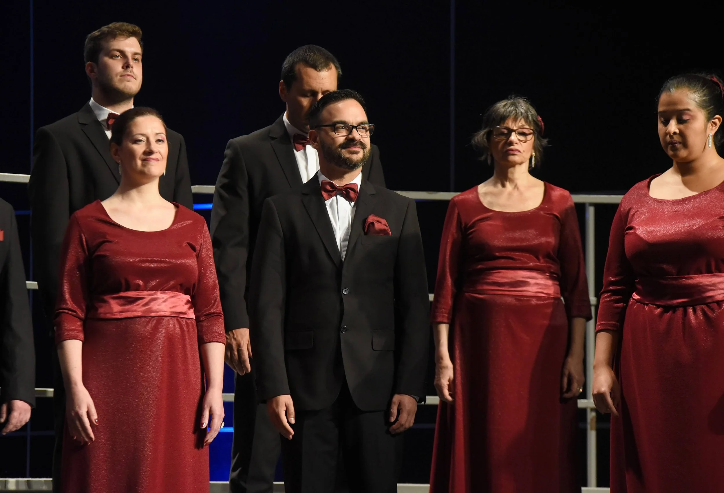 Grupo de personas en un escenario, vestido de gala con trajes negros y vestidos largos rojos, en una presentación o concierto.
