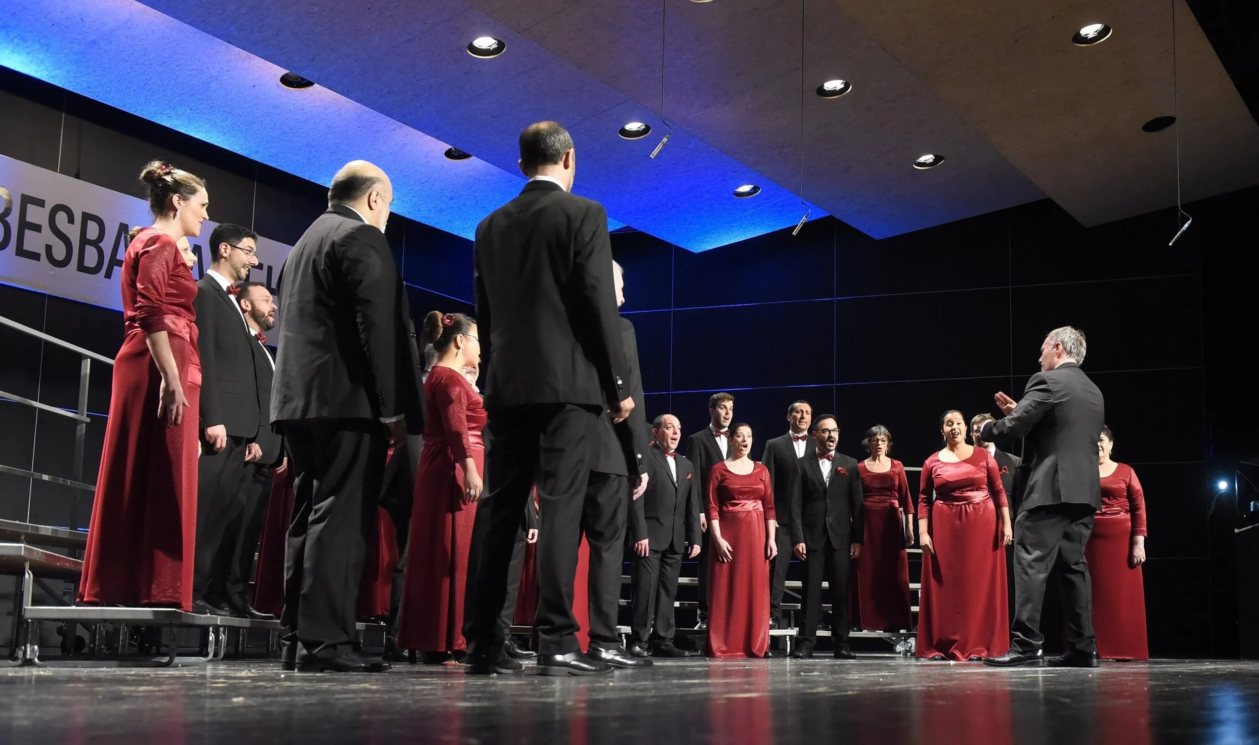 Un grupo de cantantes de coro vistiendo ropa formal, algunos en vestido largo rojo y otros en traje negro, durante un concierto en un escenario con iluminación moderna y un fondo negro y azul.