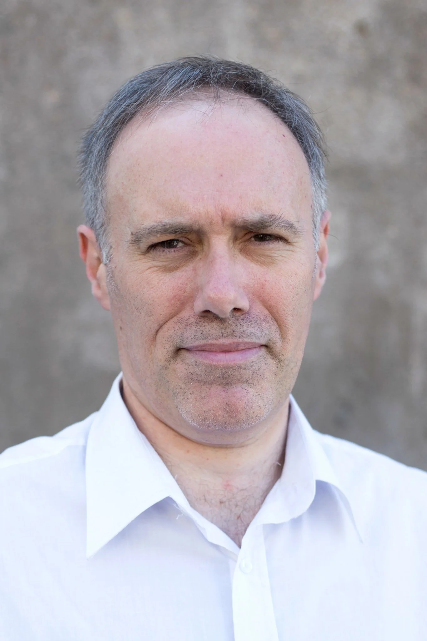 Retrato de un hombre con cabello canoso y vestido con camisa blanca, realizando una expresión facial neutral, con fondo gris.