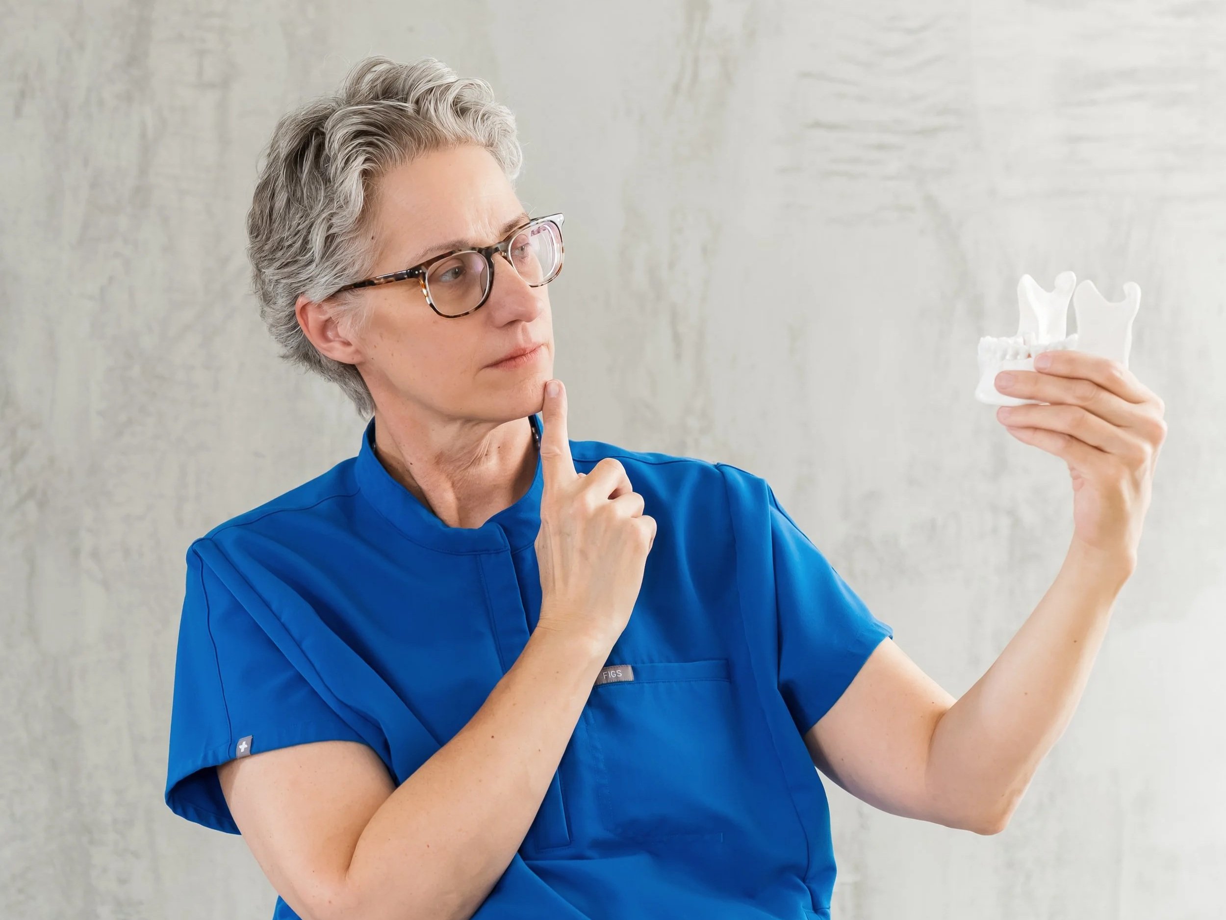 A female healthcare professional with short gray hair and glasses holds a dental model of a jaw and teeth, thoughtfully examining it while touching her chin with her other hand.