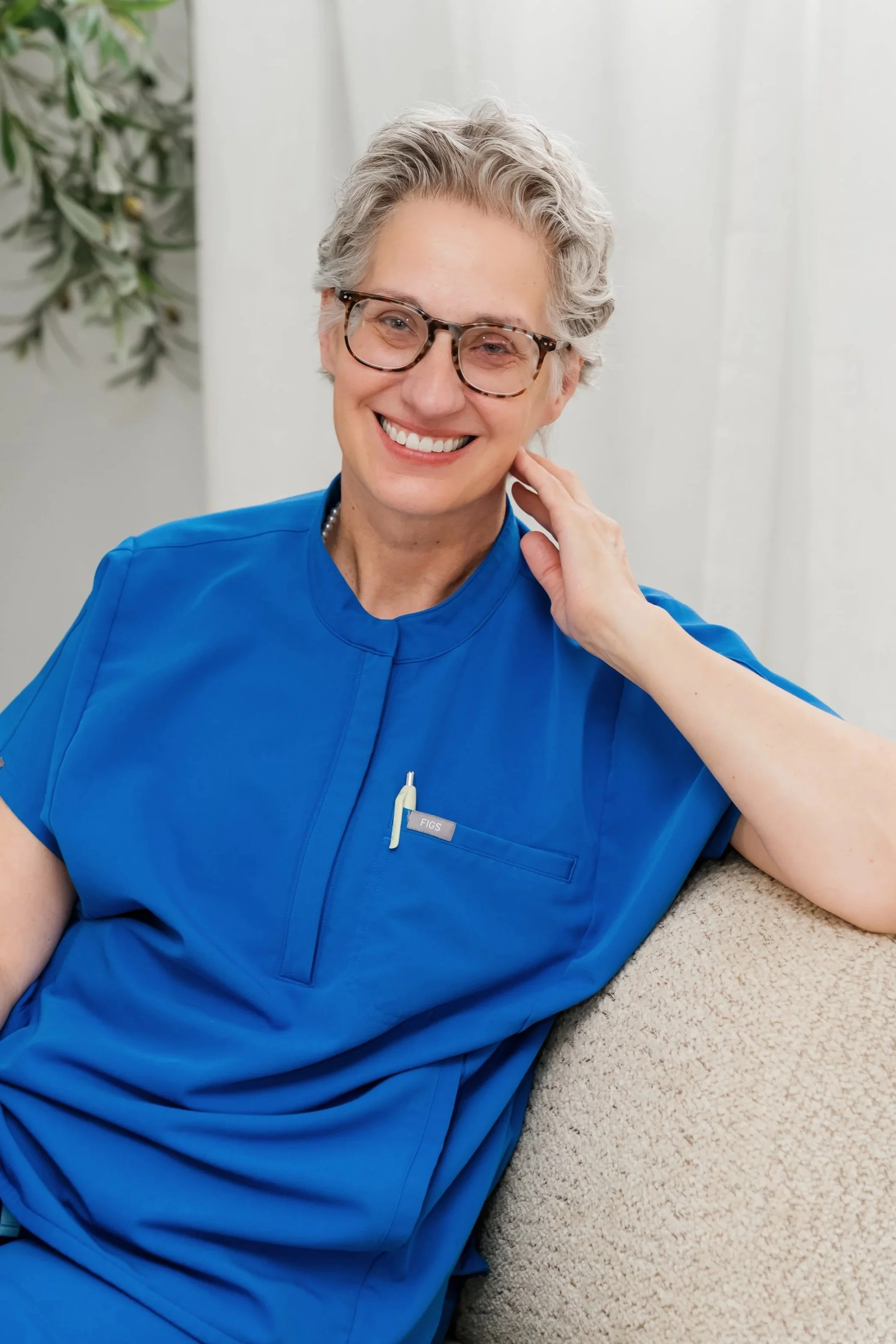 A smiling older woman with short gray hair and glasses, wearing blue scrubs, sitting on a beige couch with a white background and a plant in the background.