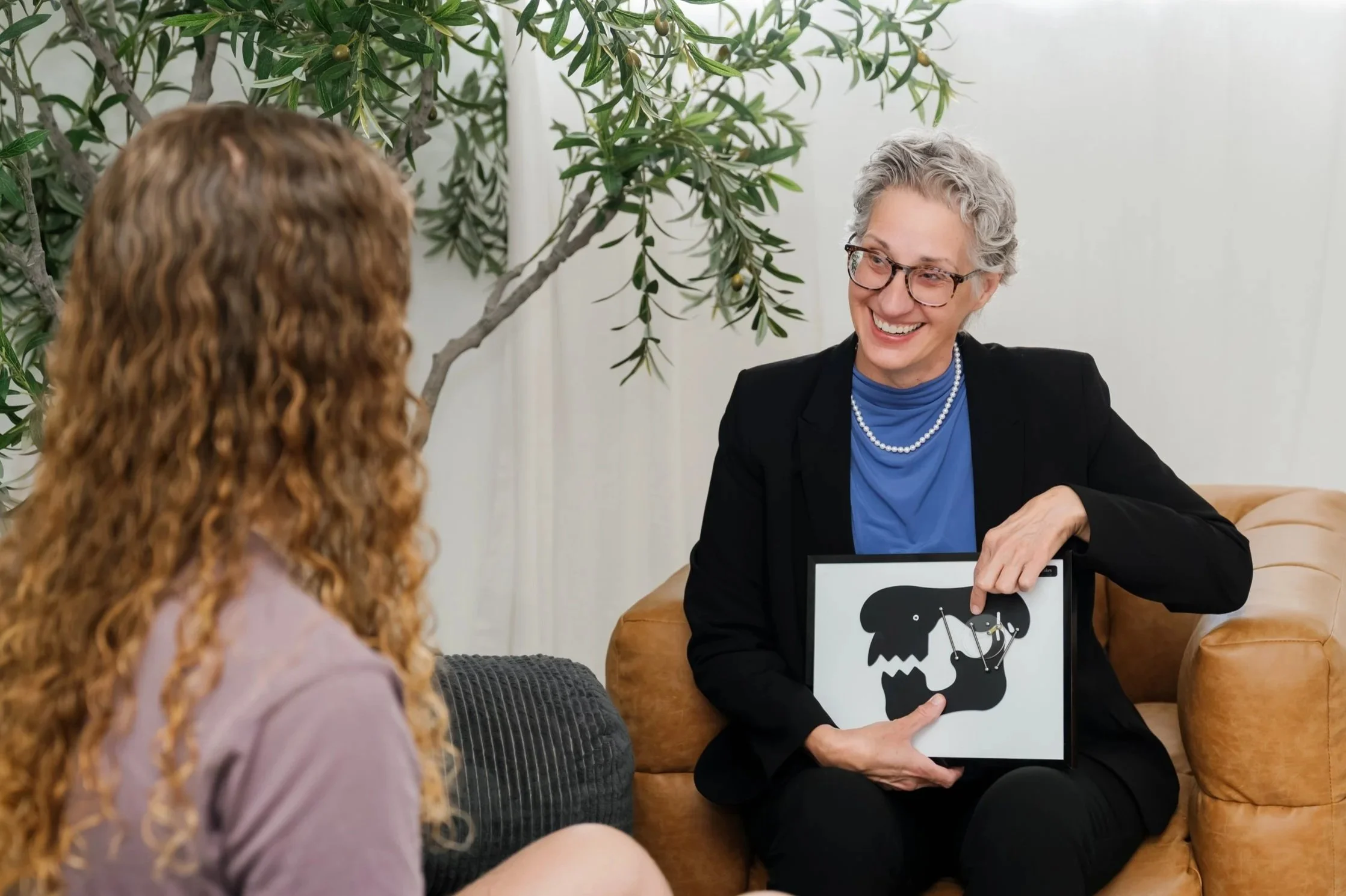 A woman with gray hair and glasses smiling, sitting on a tan sofa, holding a tablet with an abstract black and white image. She is talking to another woman with curly red hair, in a room with a large leafy plant and white walls.