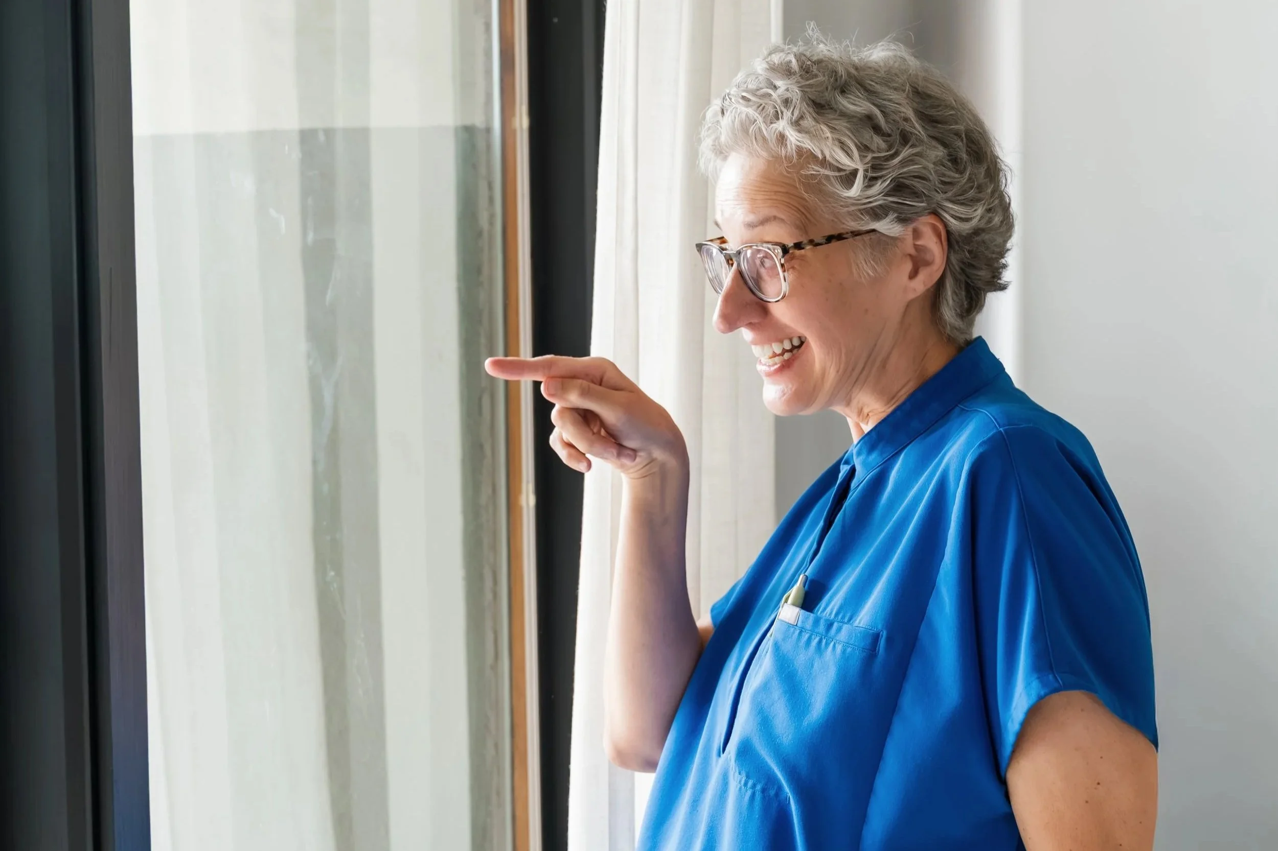 A smiling older woman with short curly gray hair and glasses, wearing blue medical scrubs, looking outside through a window.