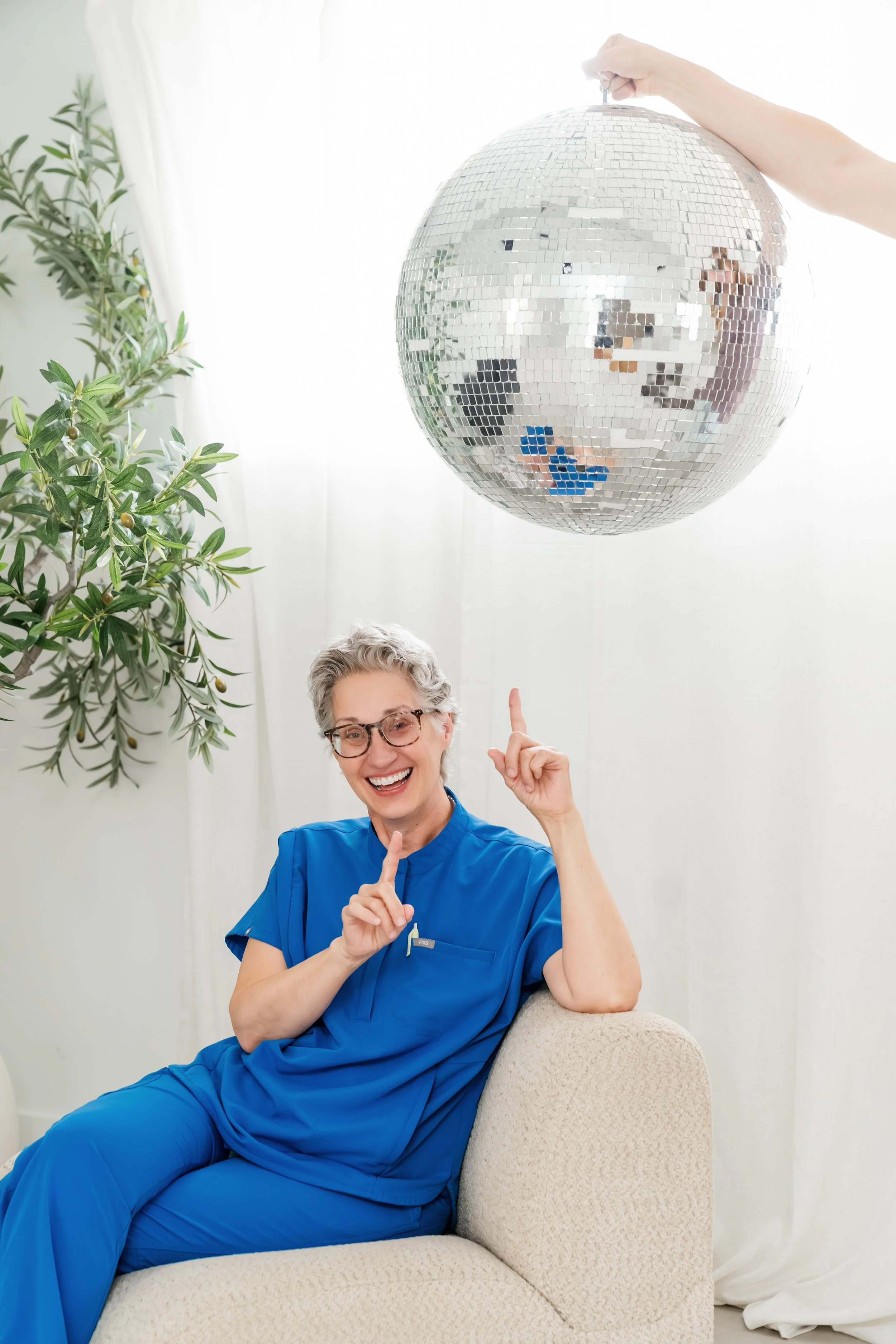 A smiling woman, Dr. Houle, in blue scrubs sitting on a beige couch, pointing upward, with a mirror ball overhead and green plants in the background.