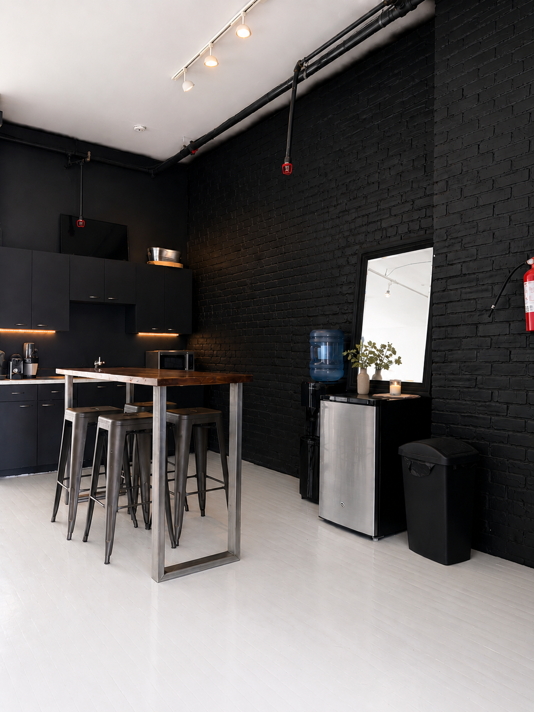 Modern kitchen with black cabinets, black brick wall, white floor, small wooden table with metal chairs, water dispenser, mirror, vase with green leaves, and a fire extinguisher.