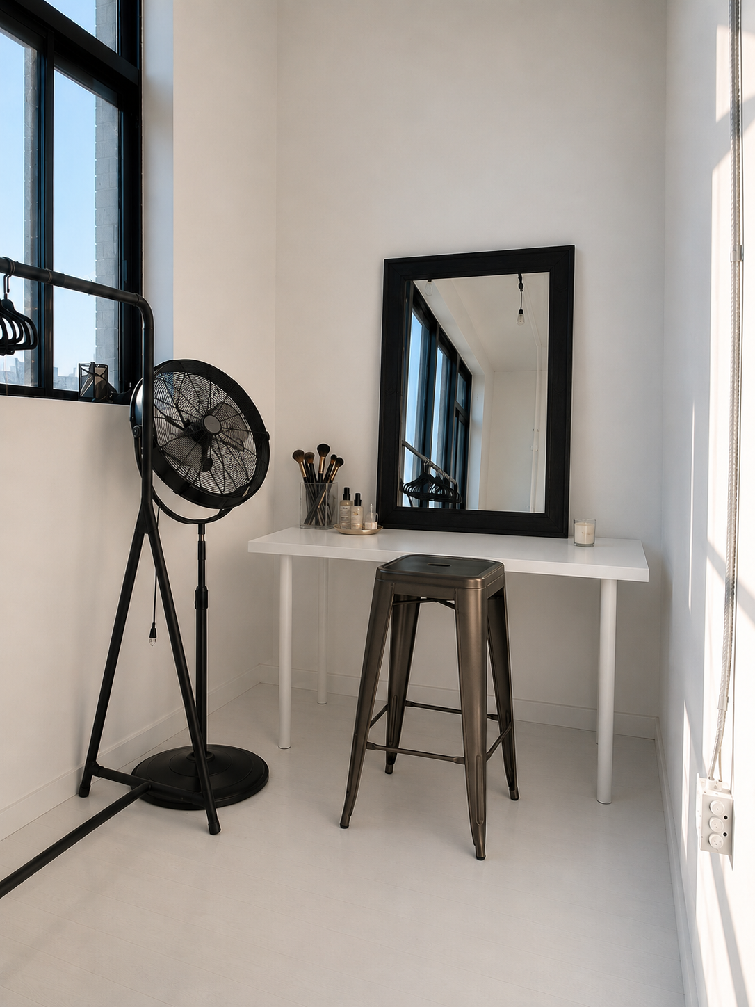 Minimalist vanity area with a white table, black-framed mirror, metallic stool, makeup brushes, and skincare products. 
