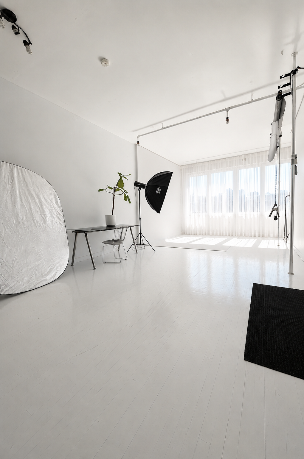 Photography studio with white walls, large window with sheer curtains, photography equipment including a softbox, tripod.