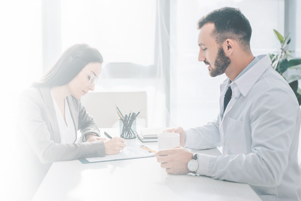 A woman in glasses writes notes at a desk while a man in a lab coat sits across from her, holding a mug, in a bright office setting.