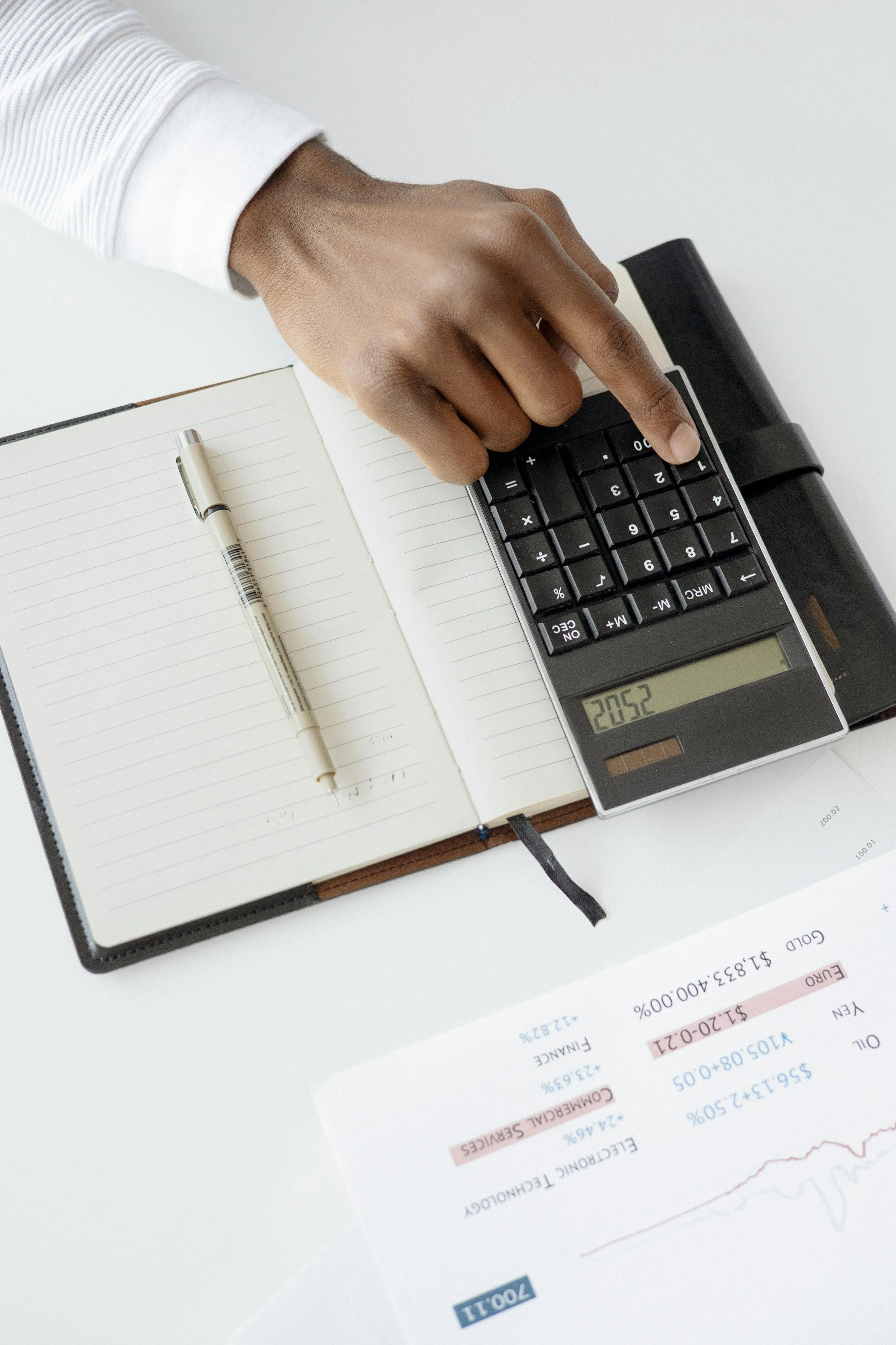 Close-up of a person's hand using a calculator on a desk. An open notebook and a pen are also on the desk, with financial documents partially visible.