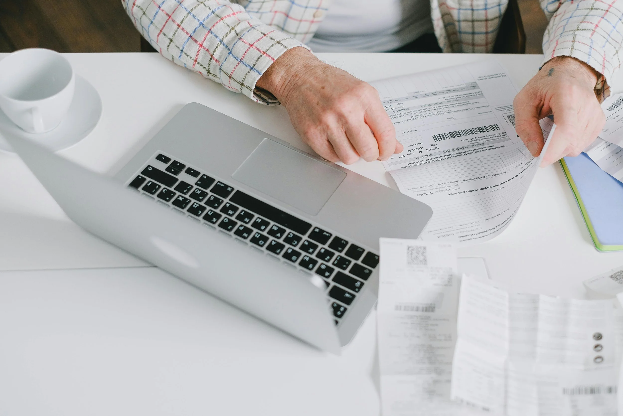 Close-up of a senior person wearing a plaid shirt, handling scattered airline tickets on a white desk next to a laptop and a white cup.