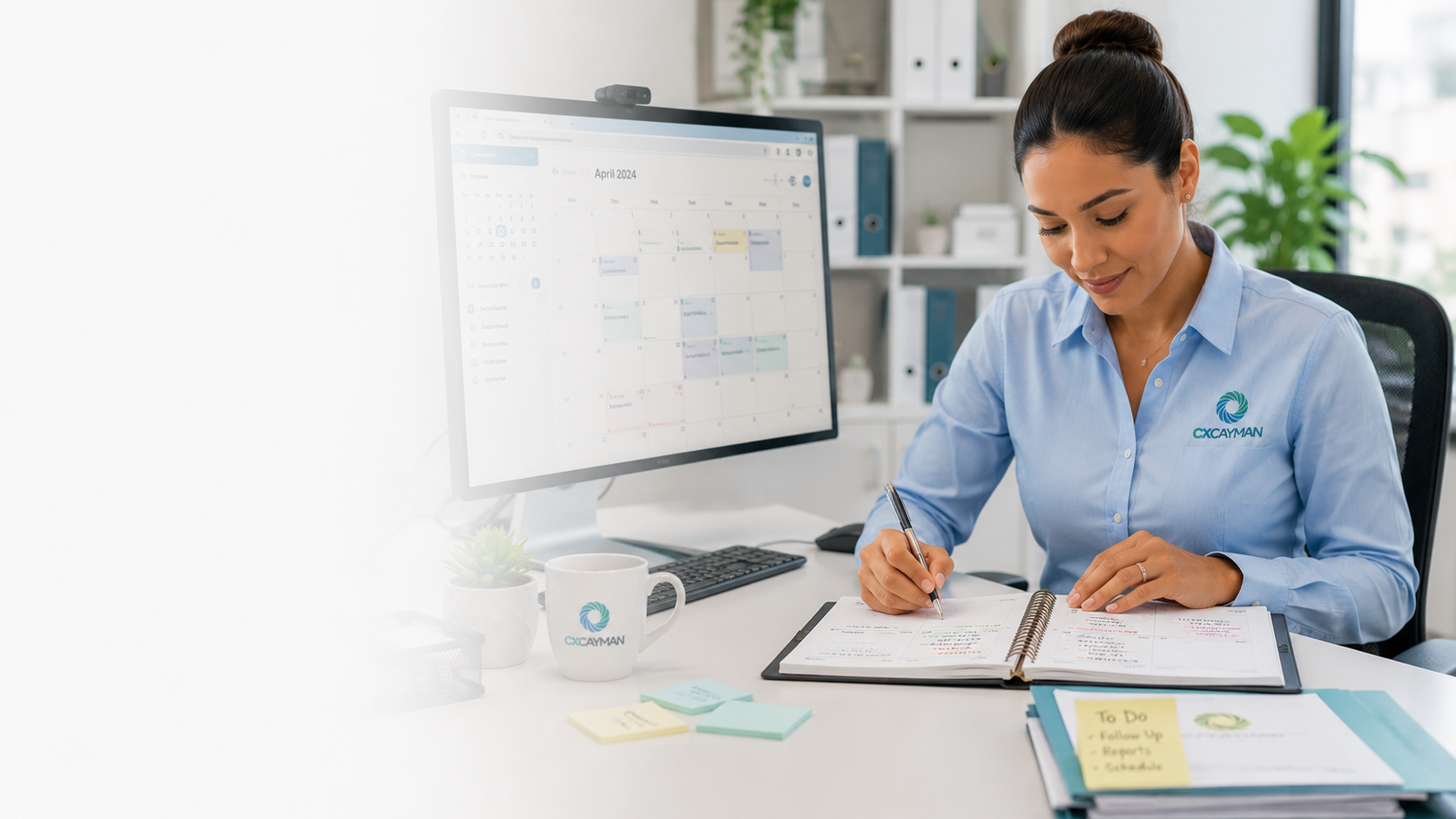 A woman in a blue shirt with a logo on it sitting at a desk, writing in a planner, with a computer monitor displaying a calendar and a coffee mug on the desk.
