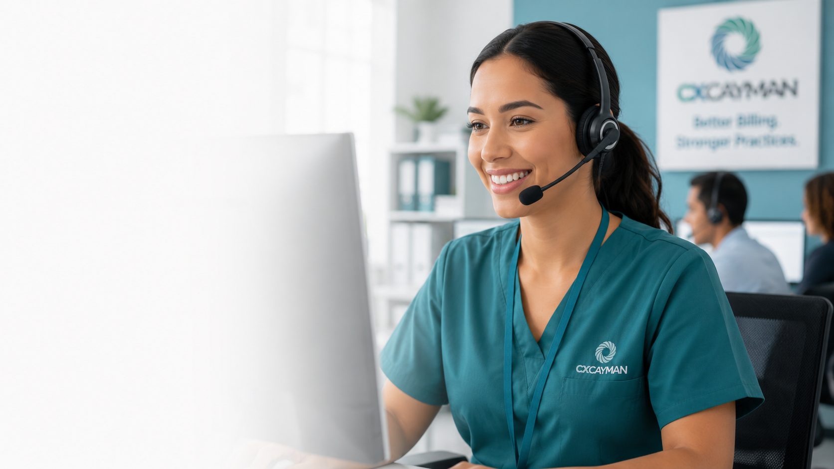 A woman wearing medical scrubs and a headset sitting at a computer in a call center or office with others working in the background.