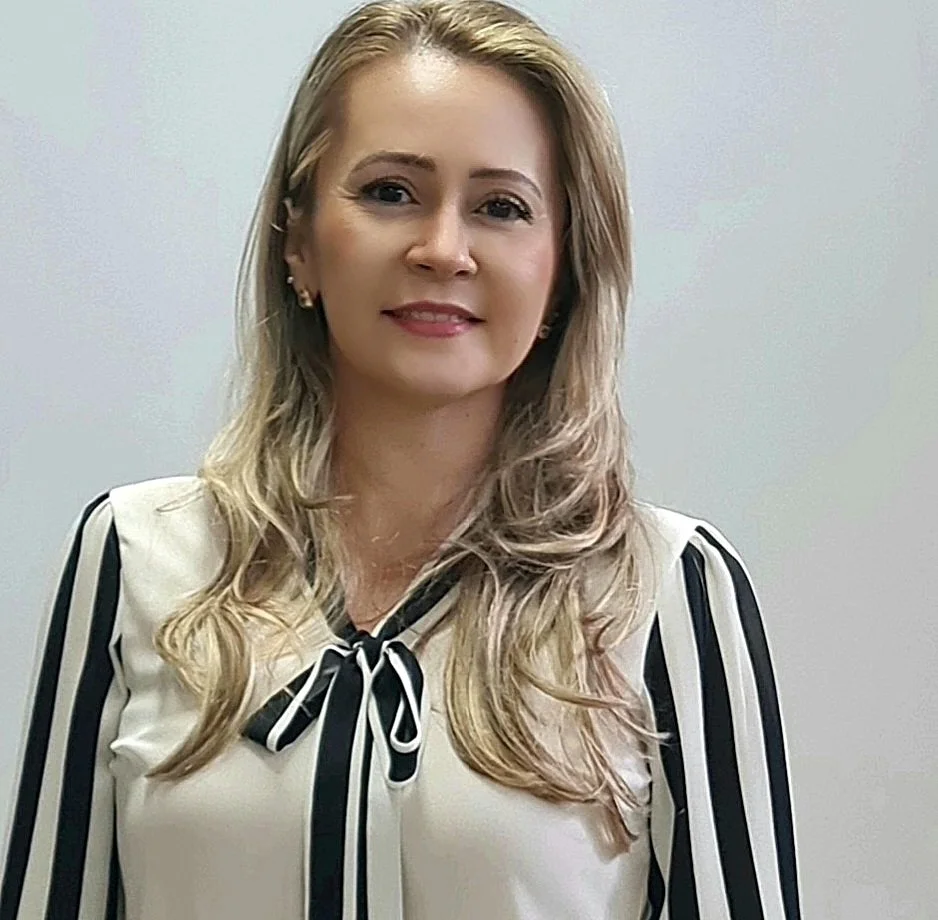 Mulher com cabelo loiro cacheado e pele clara, vestindo blusa branca com detalhes em preto, sorrindo em frente a uma parede cinza.