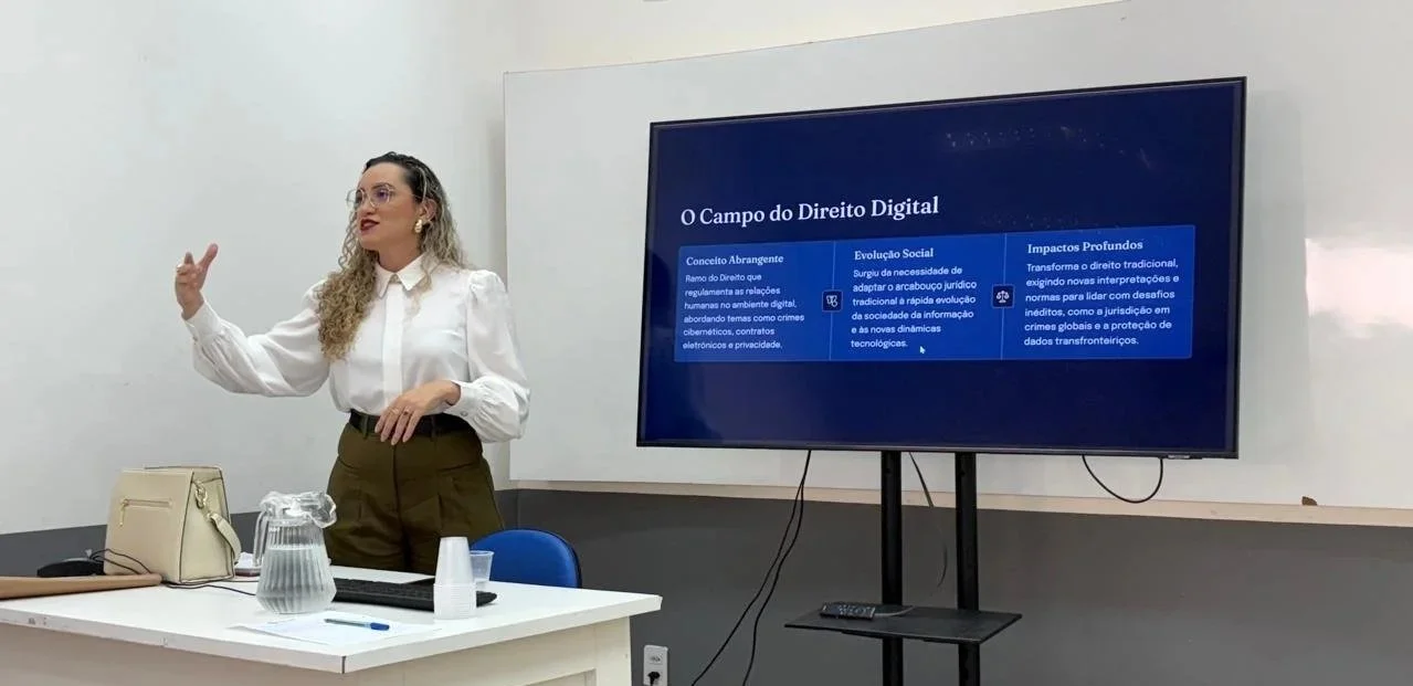 Mulher de cabelo cacheado com blusa branca e óculos durante uma apresentação, com um quadro de display digital ao seu lado.