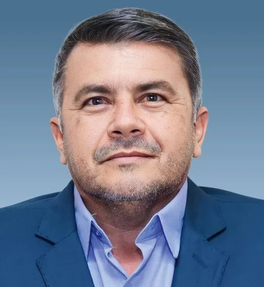 Homem de terno azul, camisa azul claro, com cabelo castanho e barba, sorrindo levemente, contra fundo cinza.