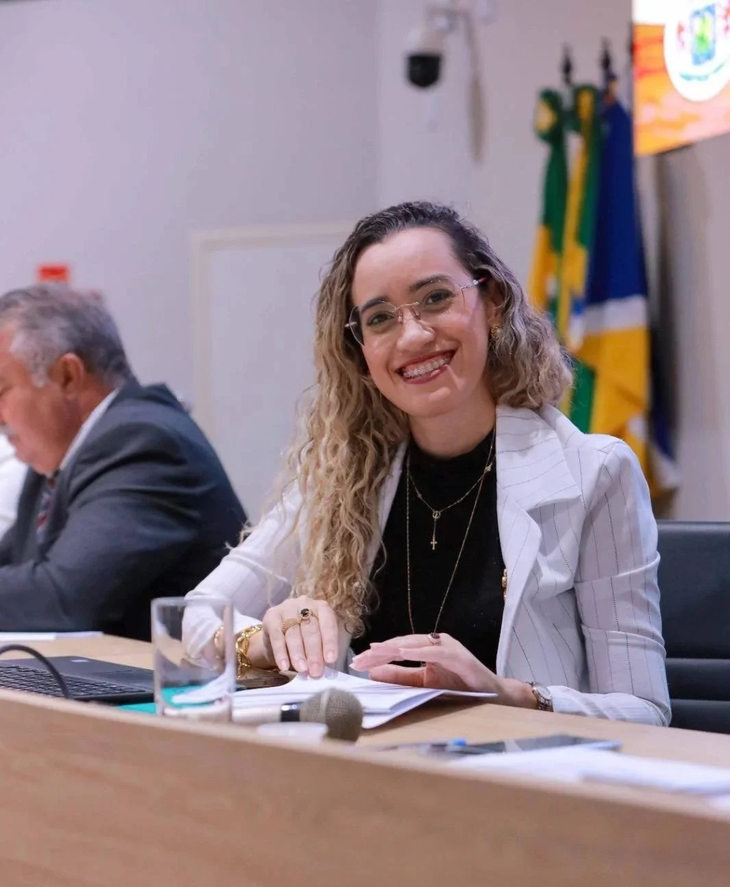 Mulher sorrindo, de cabelo cacheado e roupa branca, sentada à mesa com documentos, em ambiente formal com Bandeira do Brasil ao fundo.