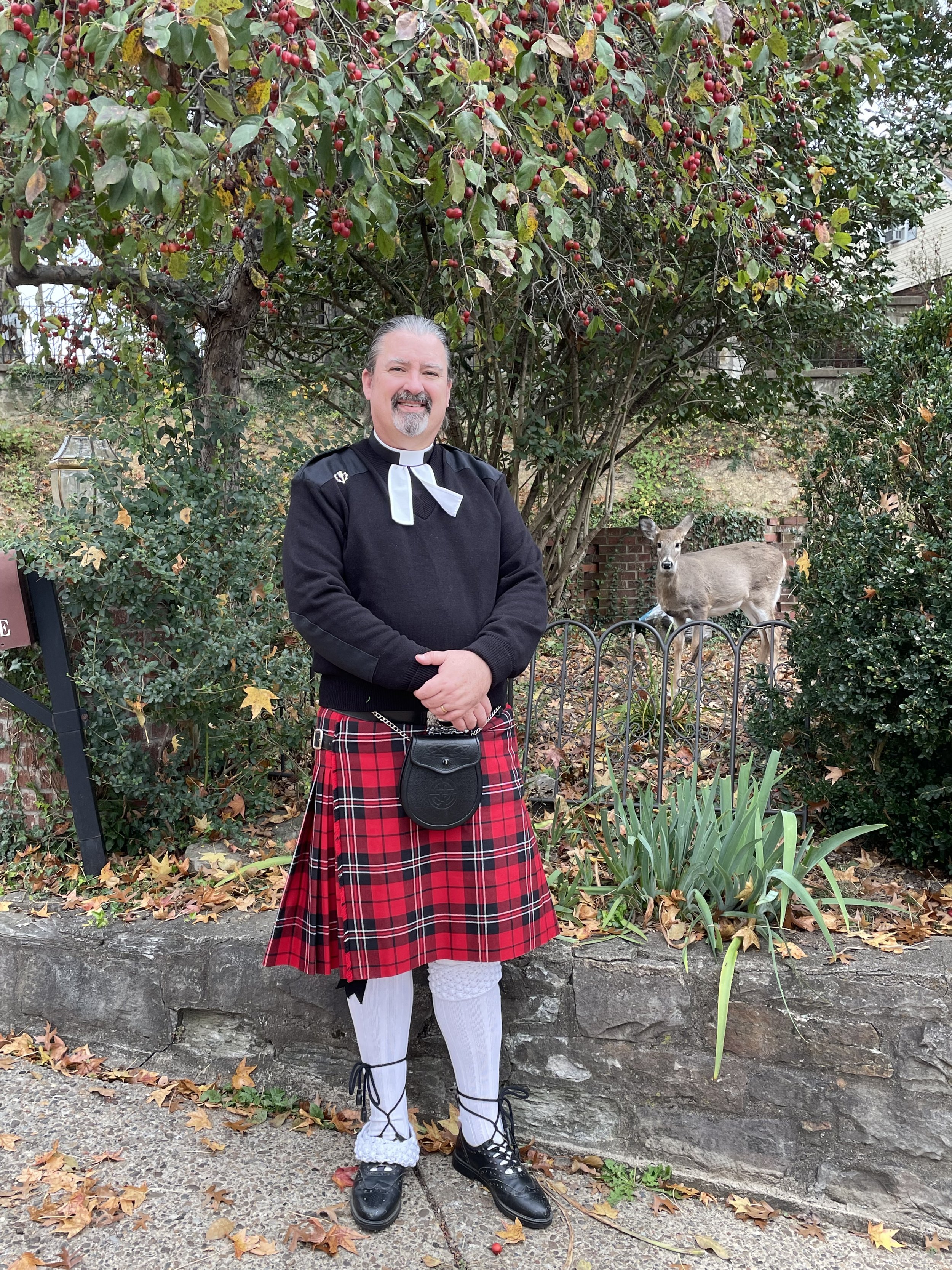 A man dressed in traditional Scottish attire, including a red tartan kilt, black dress shoes, white socks, and a black sweater, stands outdoors in front of a large bush with red berries and green leaves. He is smiling with his arms crossed, and a small deer stands nearby behind a fence. There are autumn leaves on the ground.
