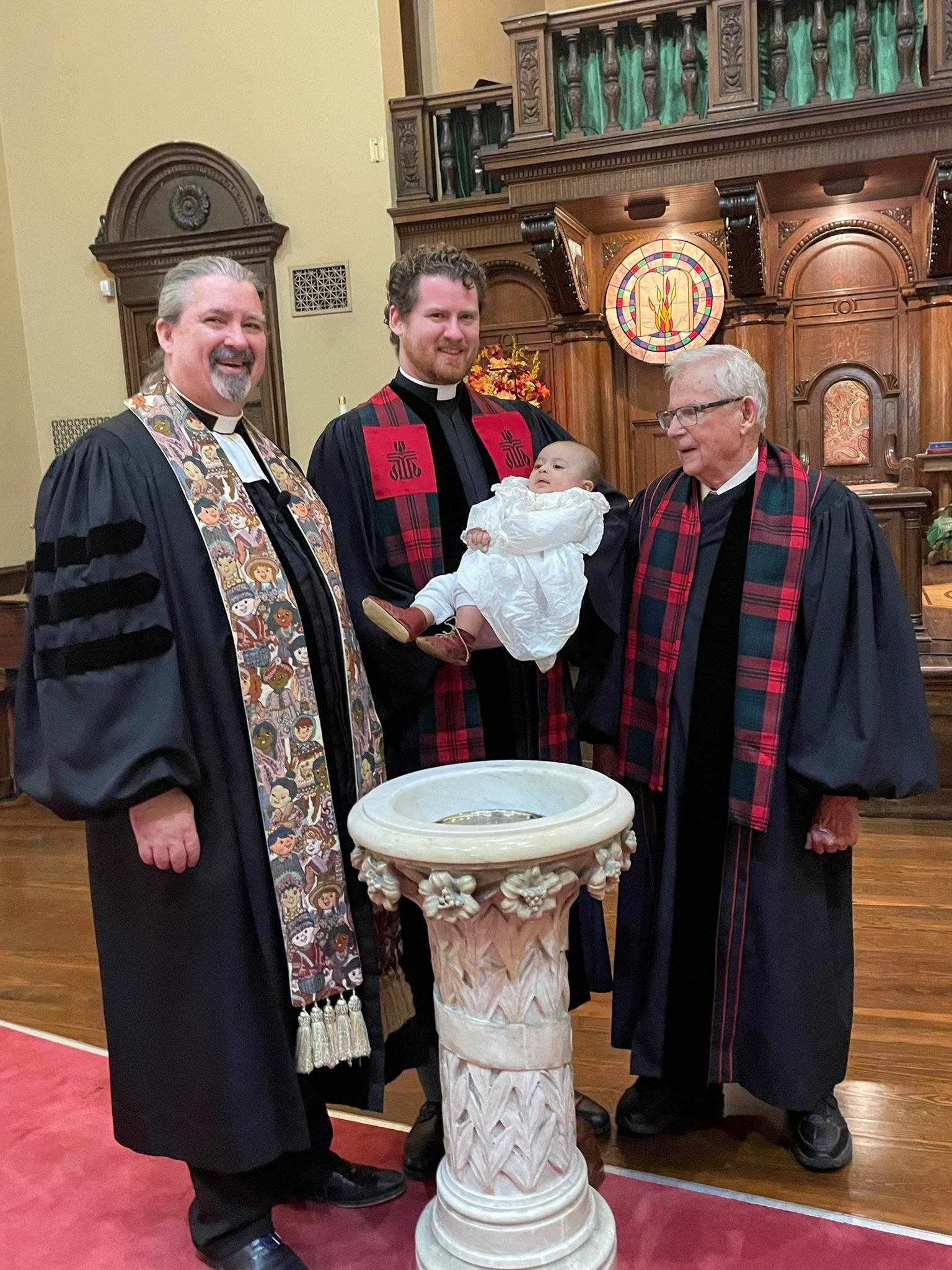 Four men, including a priest, standing inside a church around a baptismal font. One man is holding a baby dressed in white. The church has wooden decorations and stained glass windows.