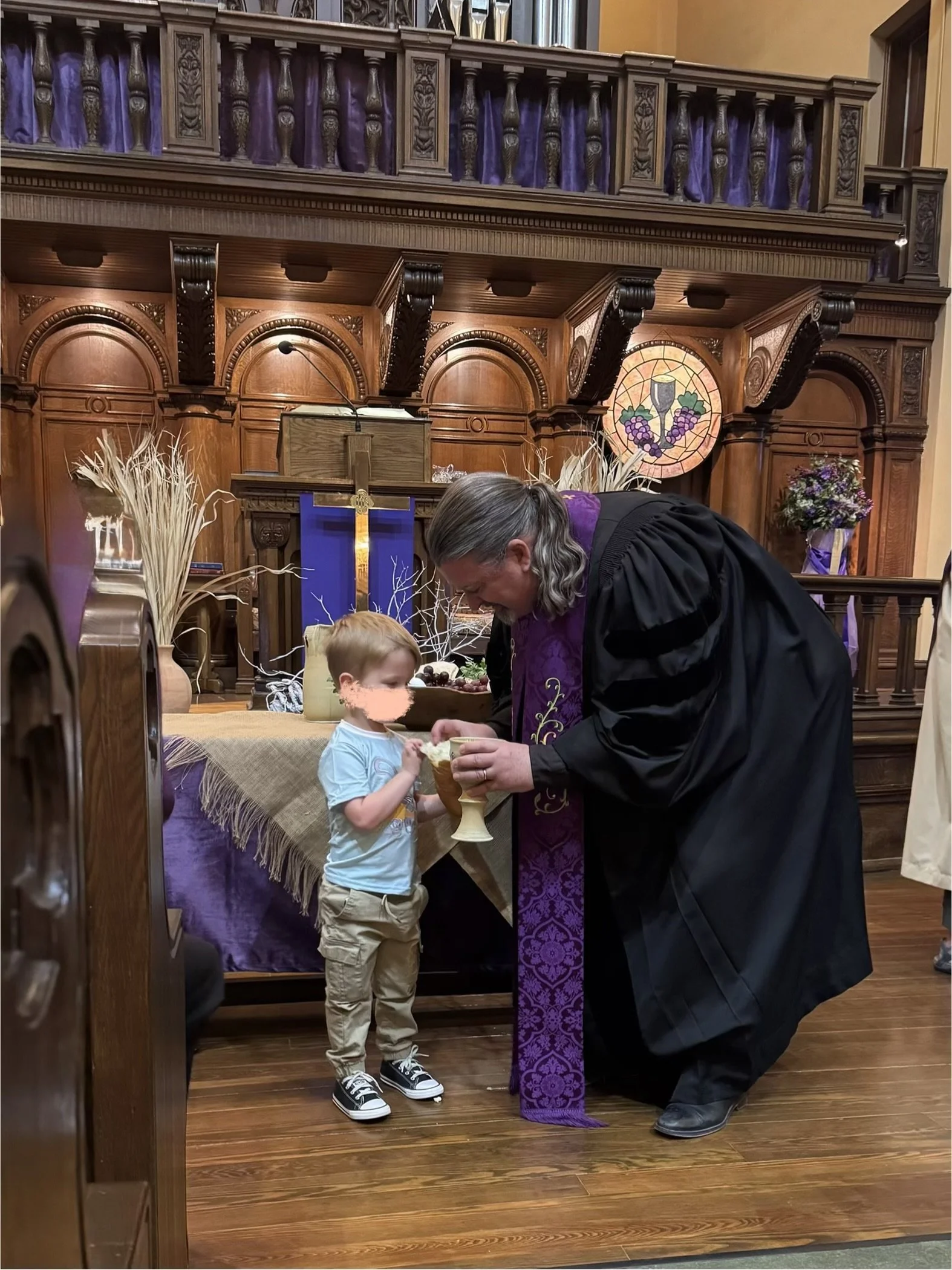 A priest with long gray hair and glasses is performing a religious ceremony with a young boy in a church. The priest is wearing a black robe with a purple stole, and the boy is dressed in a light blue shirt and tan pants. They are standing in front o
