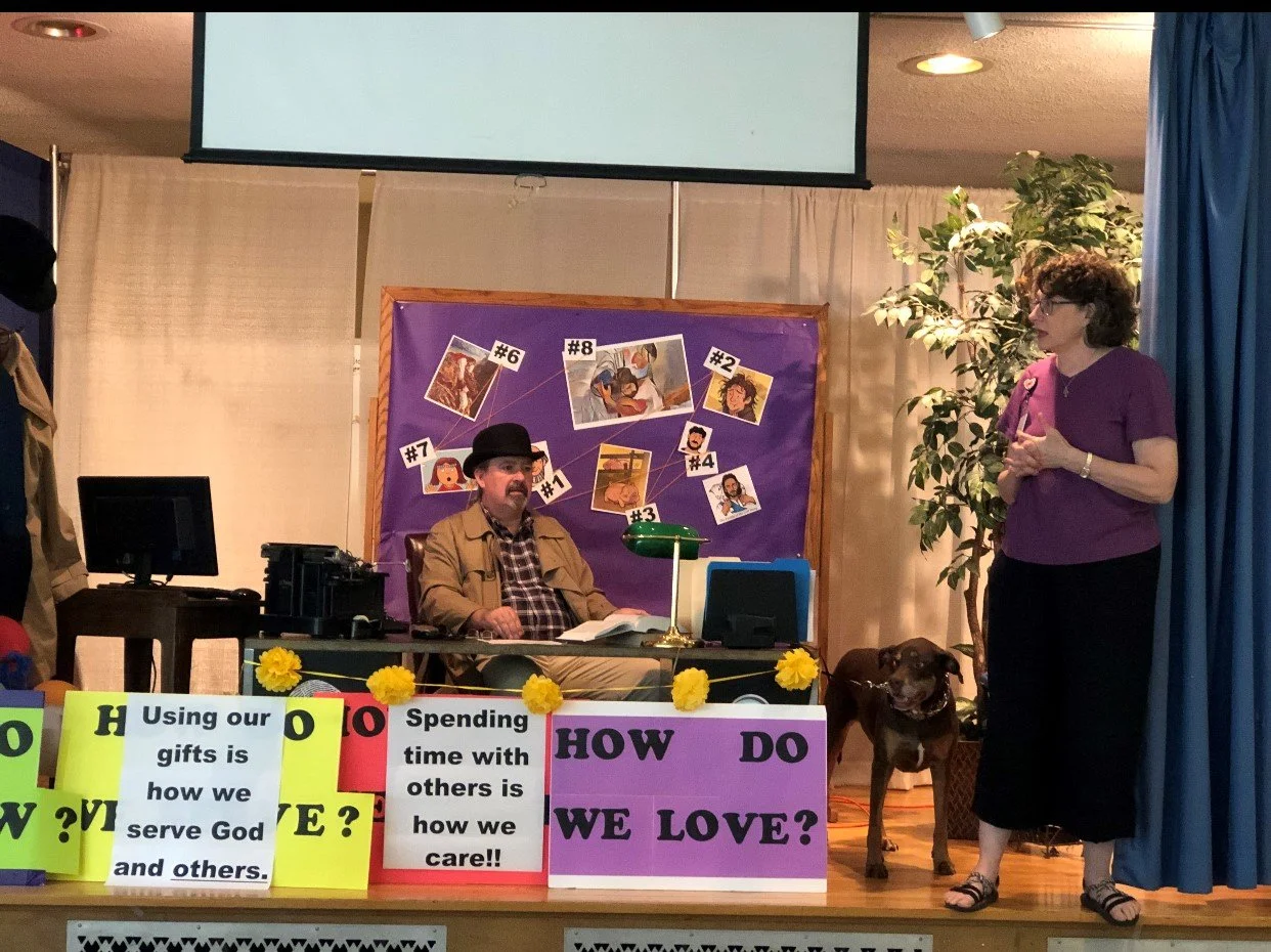 A man sitting at a desk with a woman standing to the right on a stage with a dog. The background consists of a bulletin board with pictures and numbers, and posters with questions and statements about love and service.