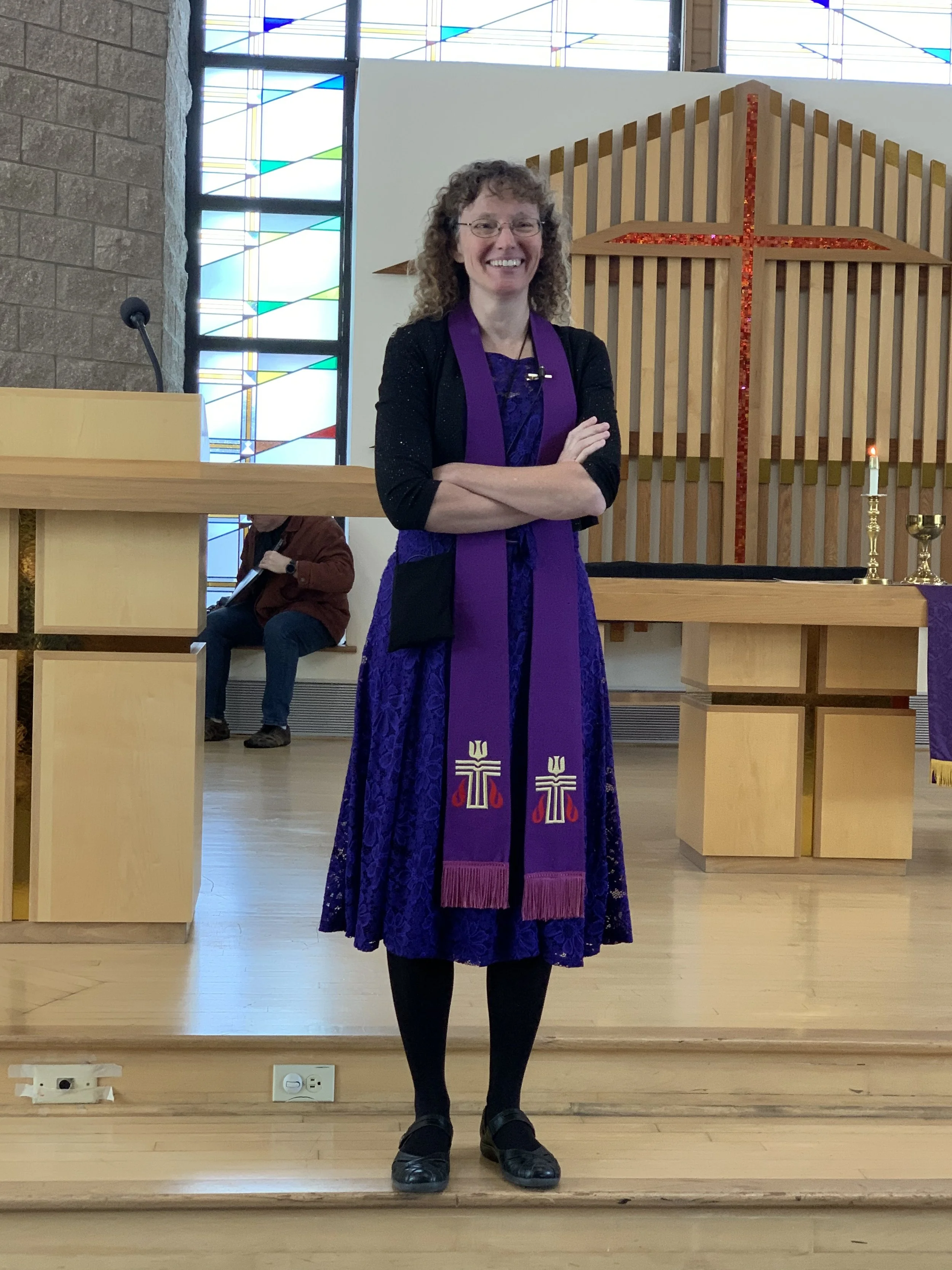 Smiling woman in purple dress and black cardigan standing with her arms crossed inside a church, with a wooden cross decorated with red glitter behind her, stained glass windows to the left, and a person sitting in the background.