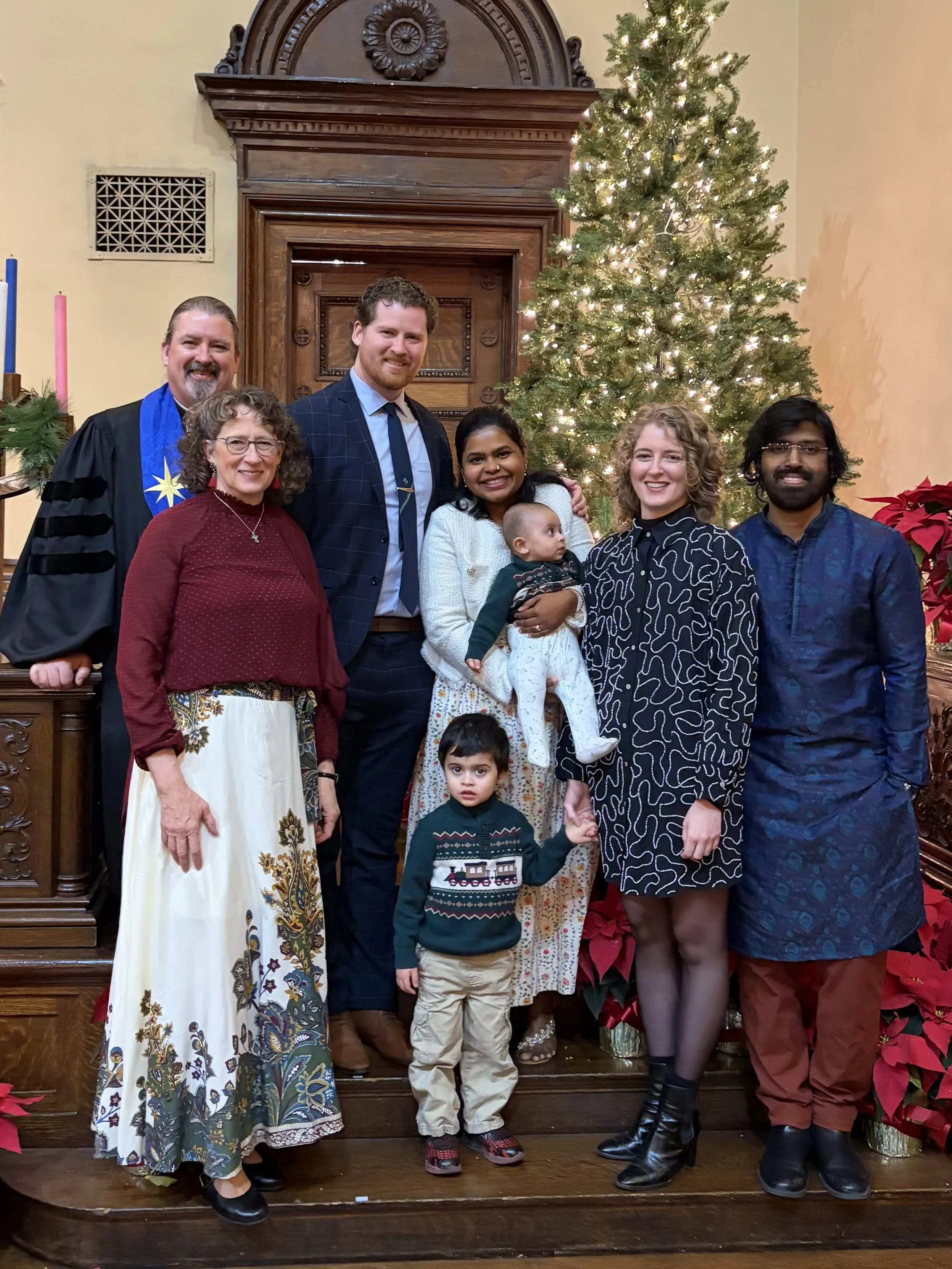 A group of seven people, including two children, posing together in front of a decorated Christmas tree and wooden backdrop. They are dressed in festive and formal attire, with a mix of older and younger adults, and smiling at the camera.