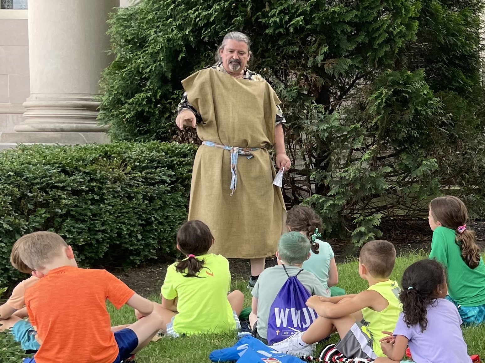 A man dressed in simple robes speaking to a group of young children outdoors, sitting on the grass with a large bush and building column in the background.