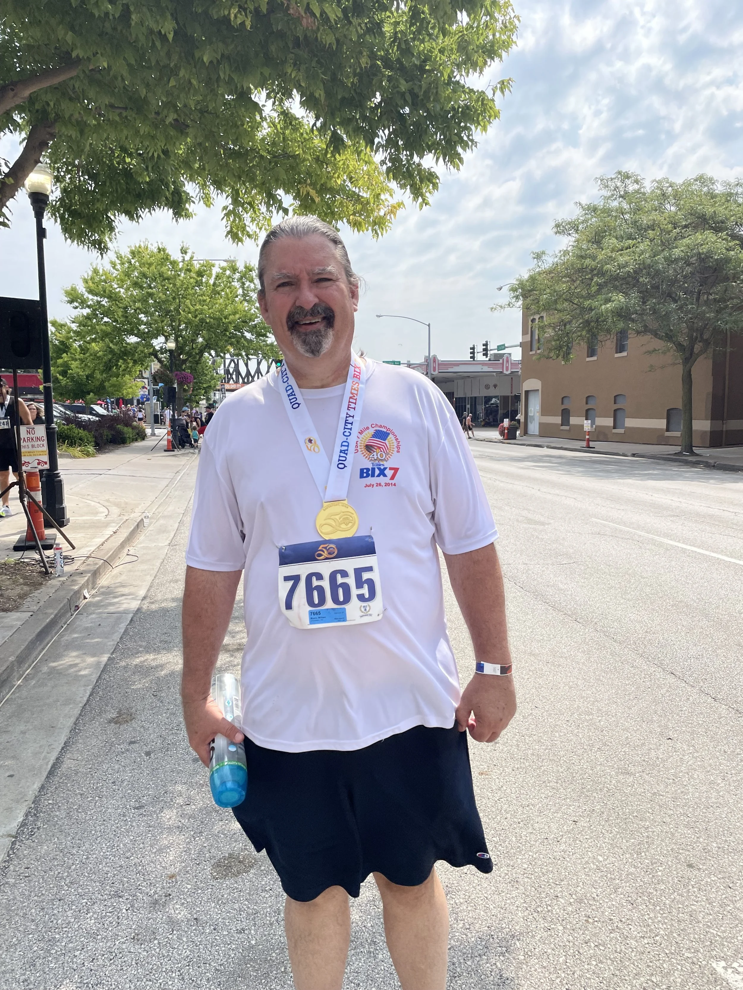Man wearing a white t-shirt and black shorts holding a water bottle, standing in a street scene with trees, buildings, and people in the background during a community event or race.