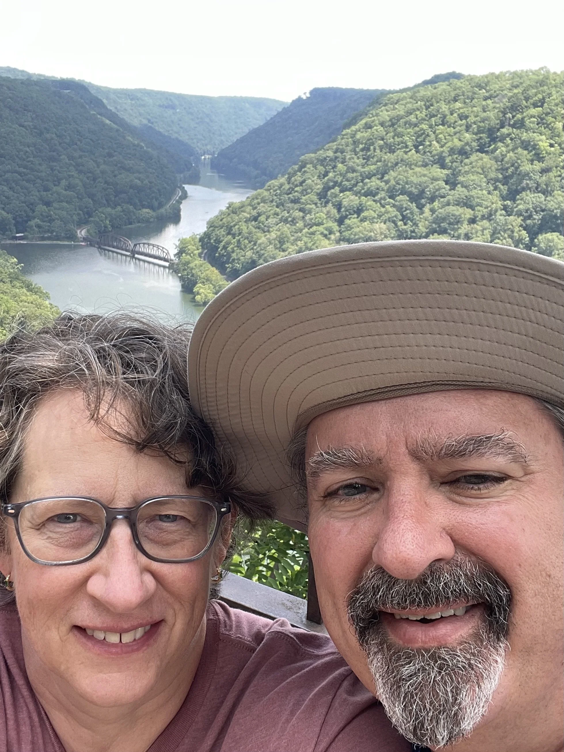 A close-up selfie of an older woman with glasses and short gray hair, and an older man with a wide-brimmed tan hat, gray beard, and short gray hair, smiling outdoors with green hills and a river in the background.