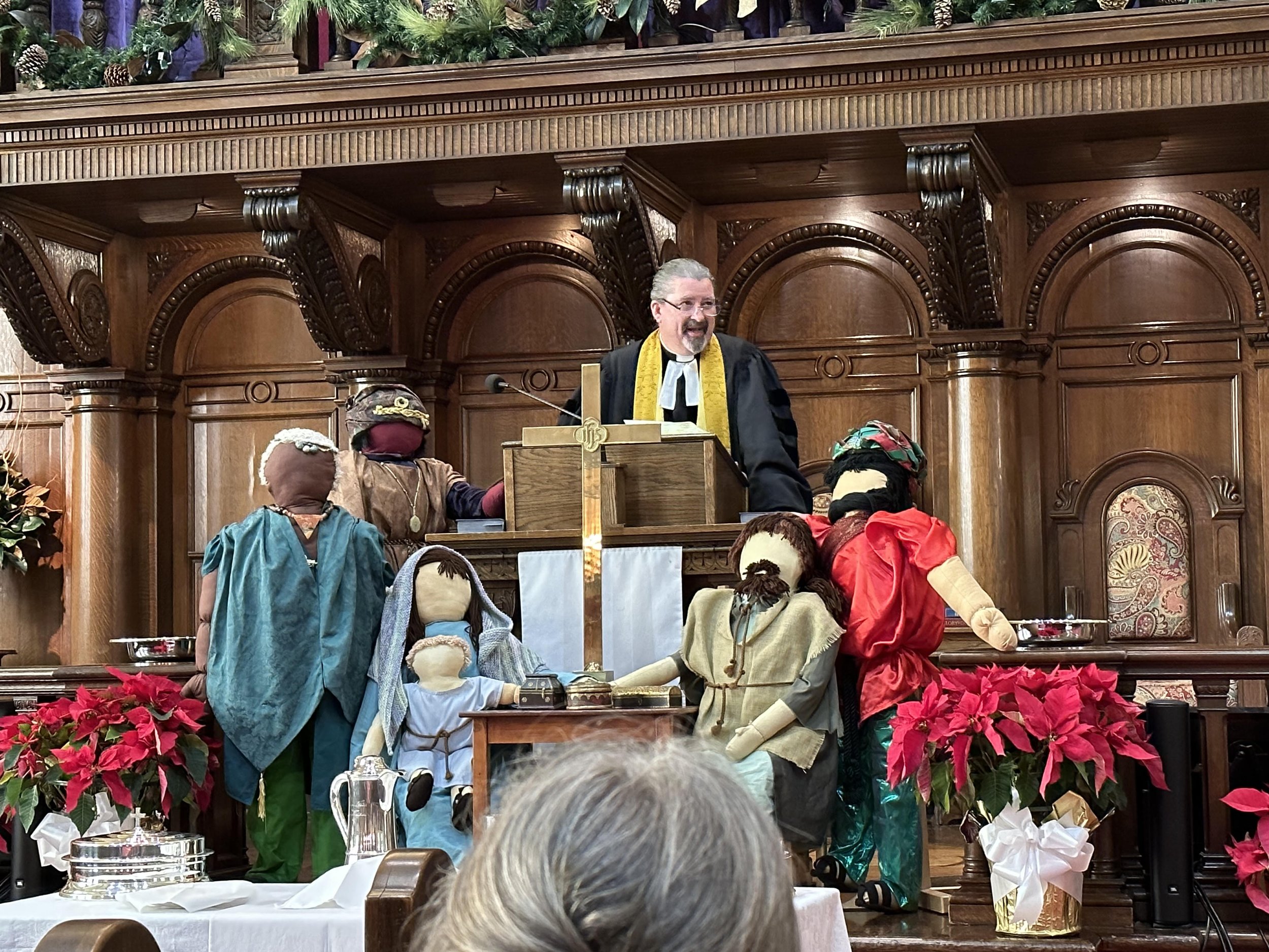 A scene from a church service featuring puppet characters and a priest. The setting is decorated with Christmas greenery and poinsettias, with a man standing behind a wooden podium. The puppet characters are dressed in colorful costumes, and the scen