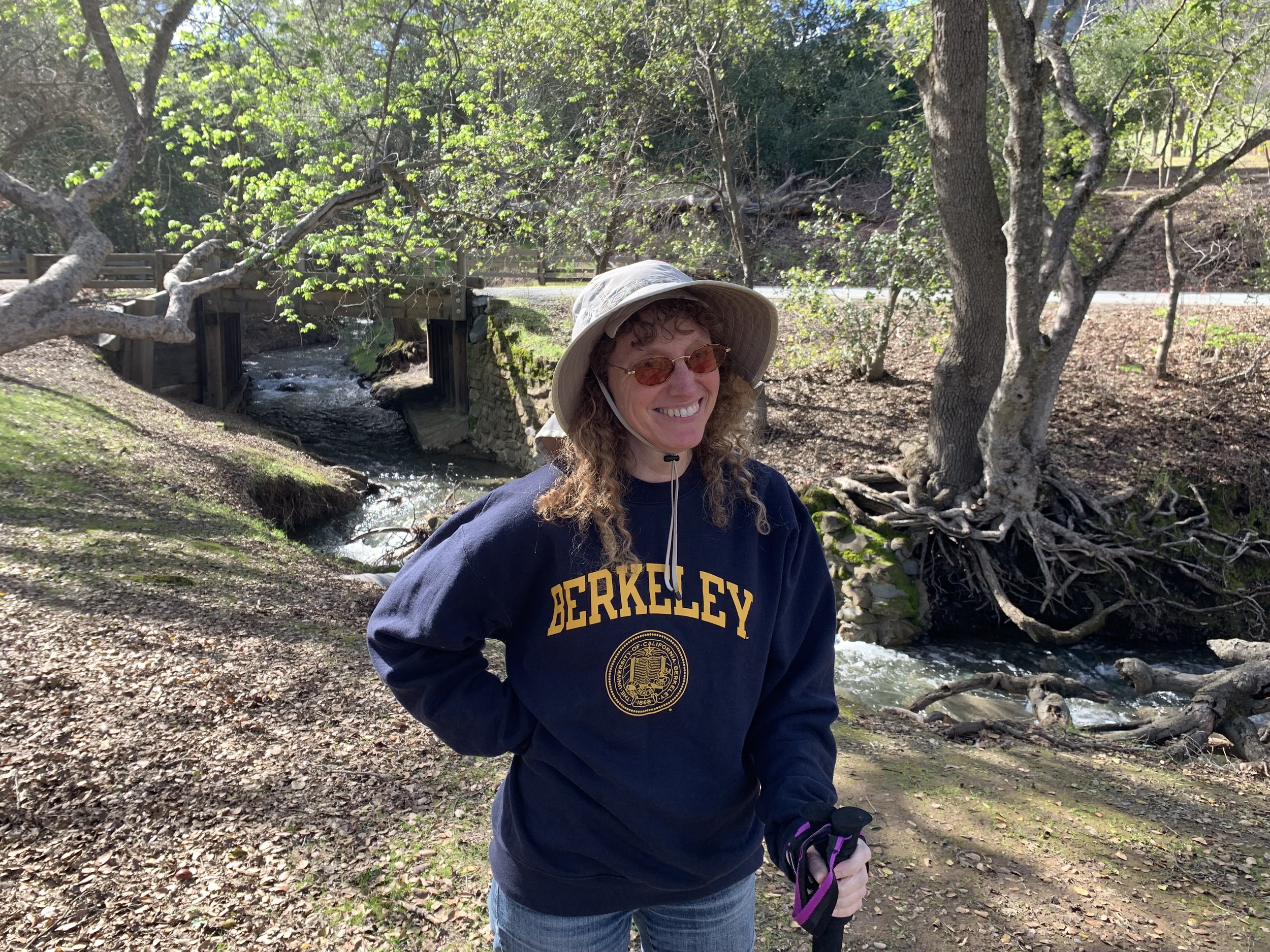 A woman with curly hair and glasses smiling outdoors in a wooded area, wearing a Berkeley sweatshirt and a wide-brimmed hat, holding a leash while standing near a stream with trees and a wooden bridge in the background.