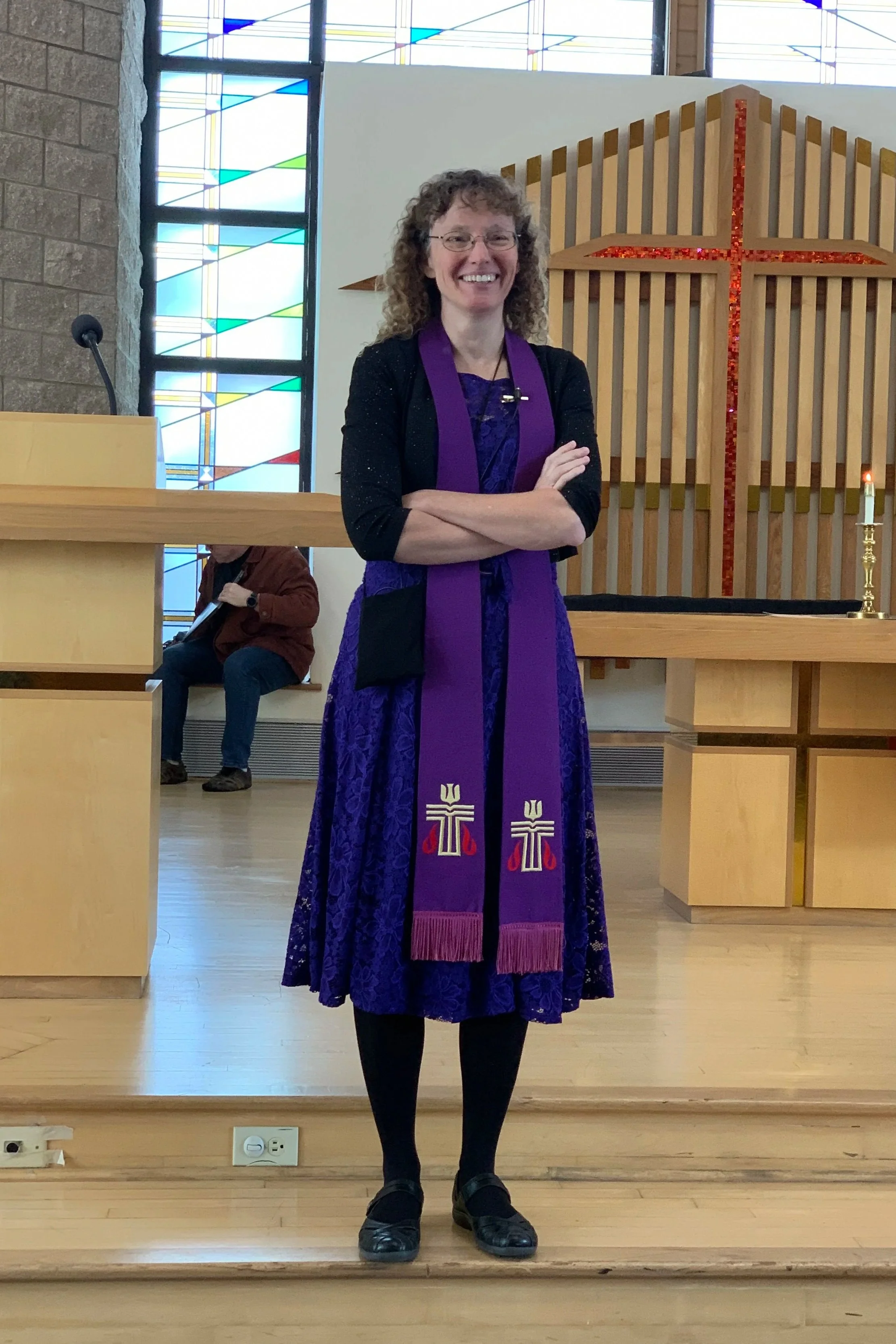 A woman wearing a purple dress and a purple stole with crosses, standing at the front of a church sanctuary with her arms crossed and smiling. Behind her are stained glass windows and a large wooden cross with red accents.