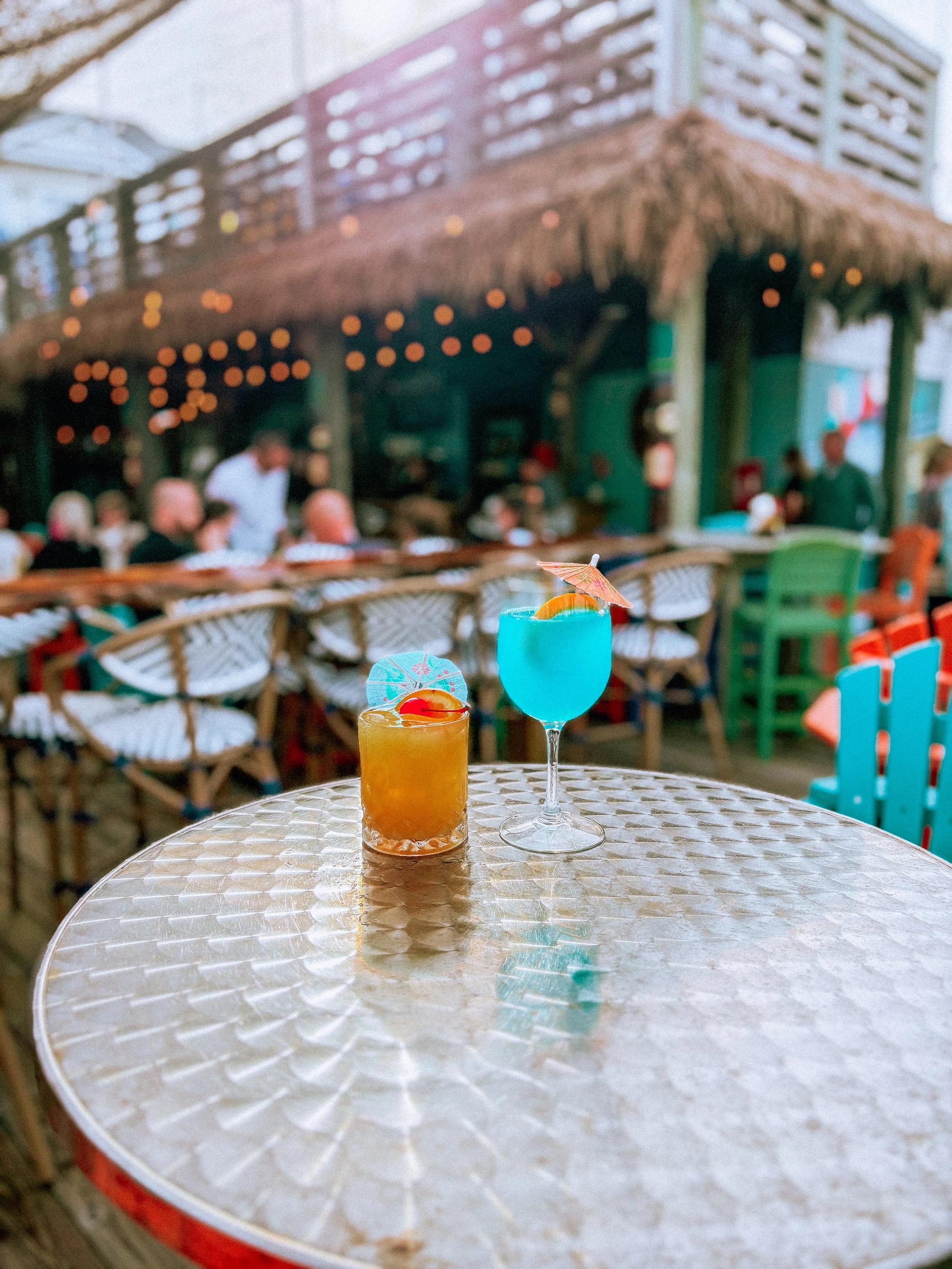 Close-up of a table with two colorful cocktails, one orange with a small paper umbrella and cherry, and a blue one with a small palm tree umbrella, in an outdoor bar or restaurant setting with people in the background.