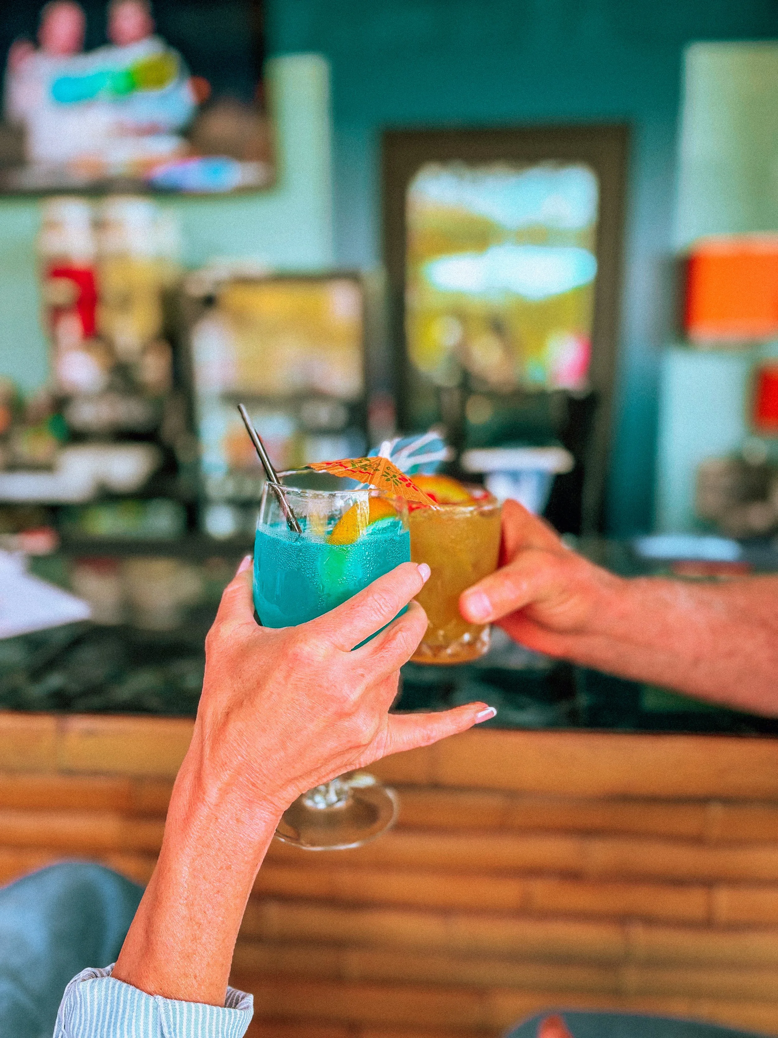 Two people clinking glasses filled with colorful cocktails, with a blurred background of a cozy, brightly decorated room.