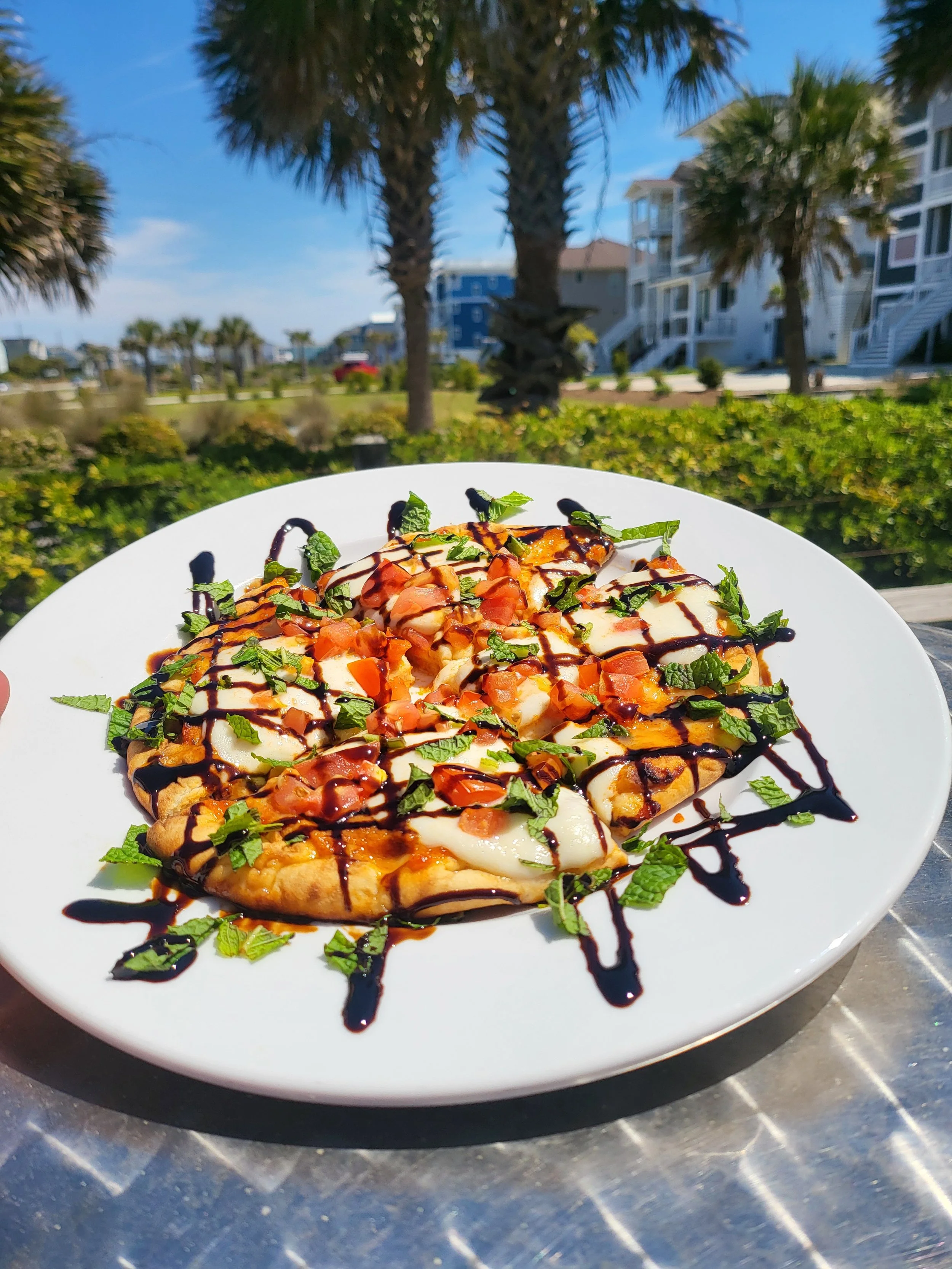 A plate of pizza topped with chopped tomatoes, fresh basil, and a drizzle of balsamic glaze, set outdoors with palm trees and a blue sky in the background.
