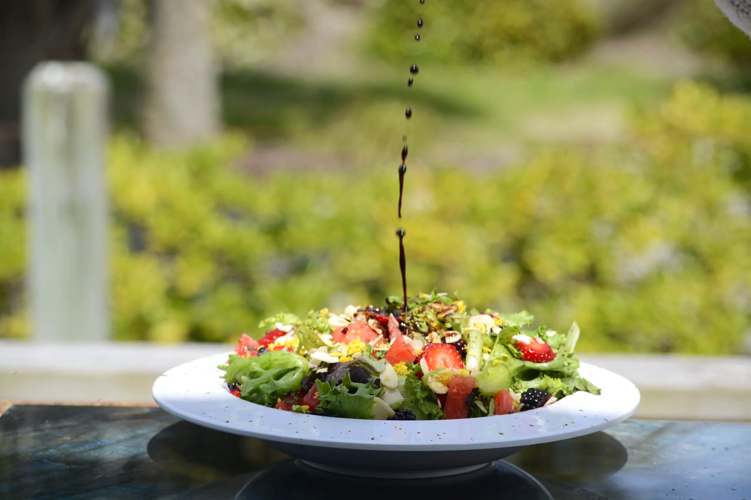 Fresh garden salad topped with a dark balsamic glaze or dressing, served on a white plate outdoors with a blurred green background.