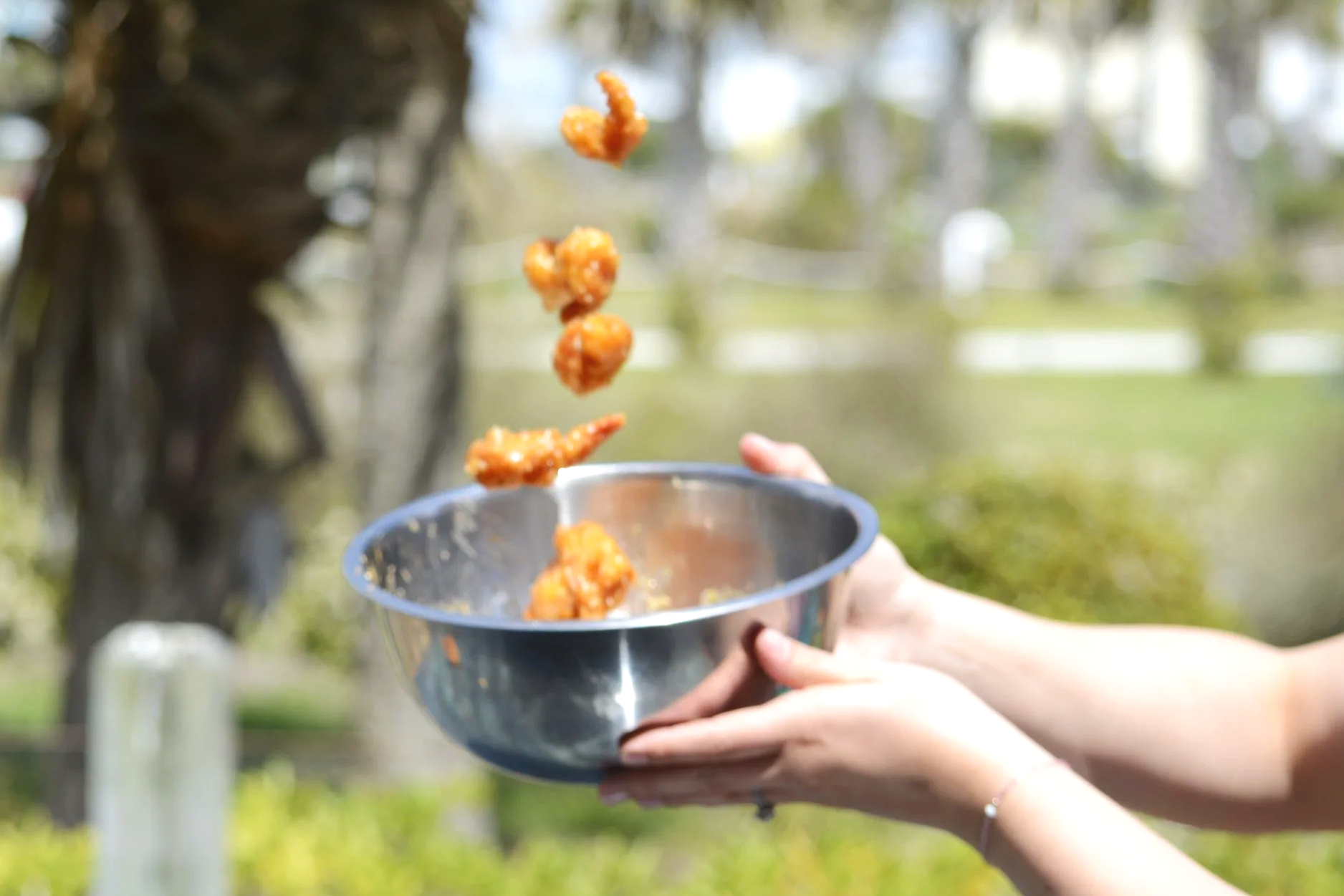Person holding a bowl tossing firecracker shrimp pieces outdoors on a sunny day.