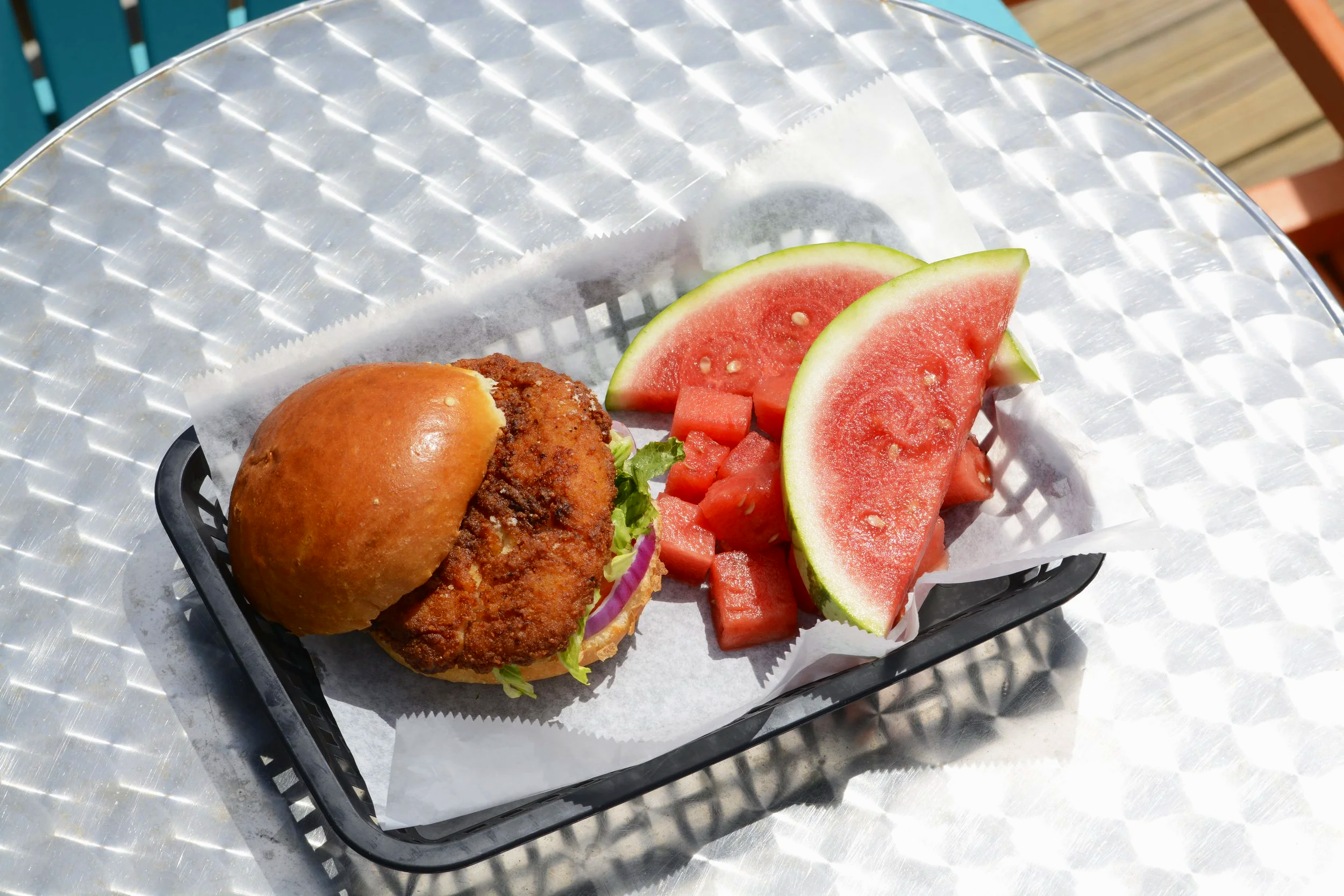 Fried chicken sandwich with lettuce and pickles, served with sliced watermelon and diced watermelon on a black tray.