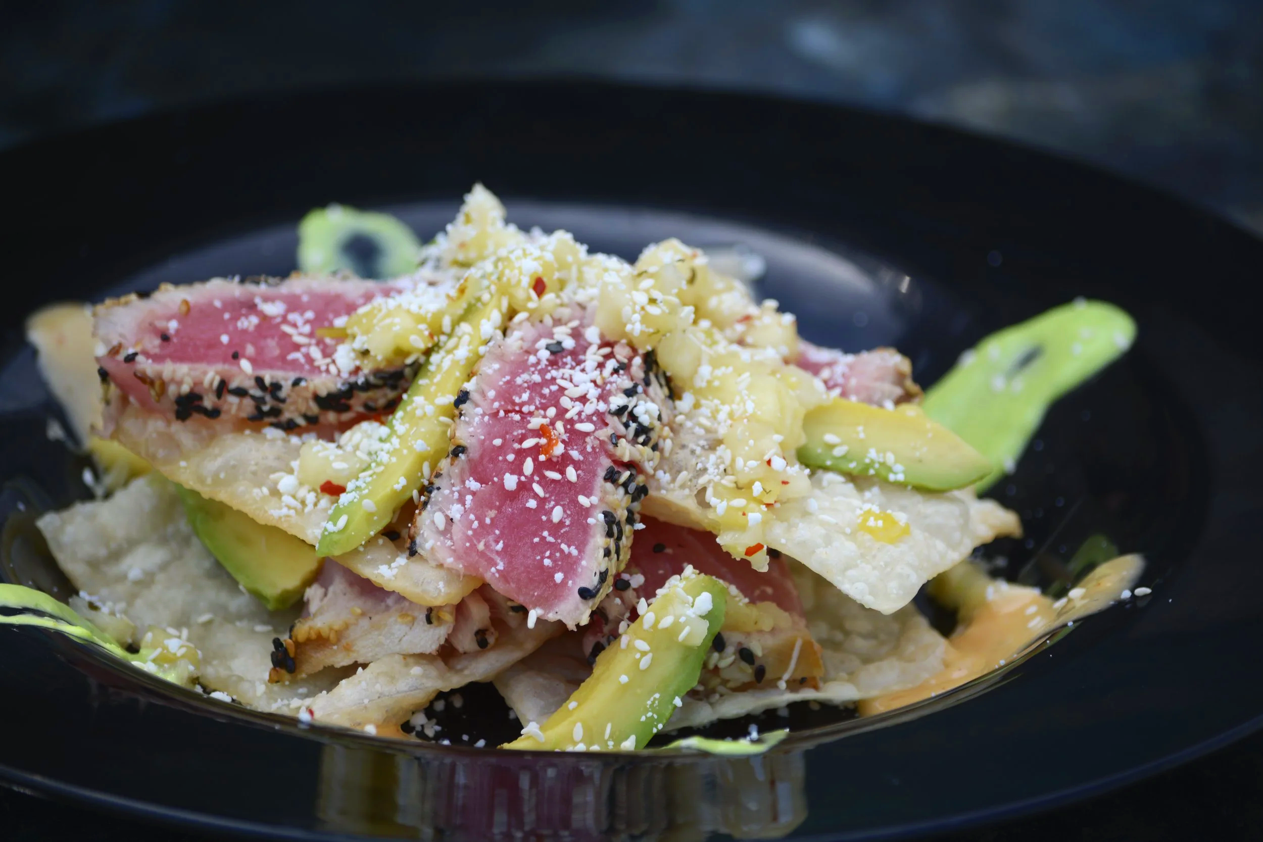 Close-up of a plate of seared tuna sashimi with sesame seeds, served with sliced avocado on a black plate.