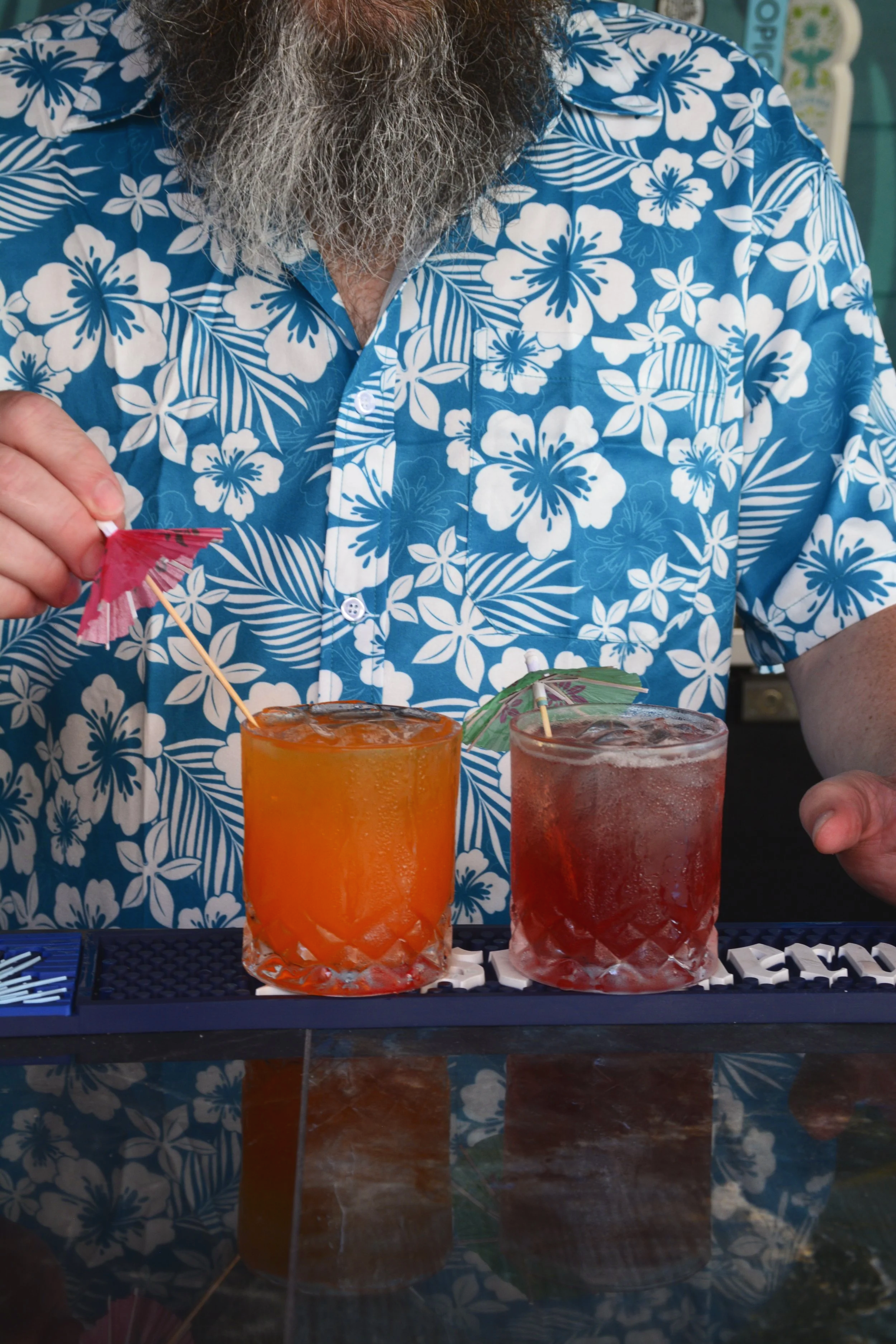 Close-up of a person with a gray beard wearing a blue Hawaiian shirt with white floral patterns. The person is preparing two tropical cocktails with umbrellas and has various bar accessories on the counter, which has a reflective surface.