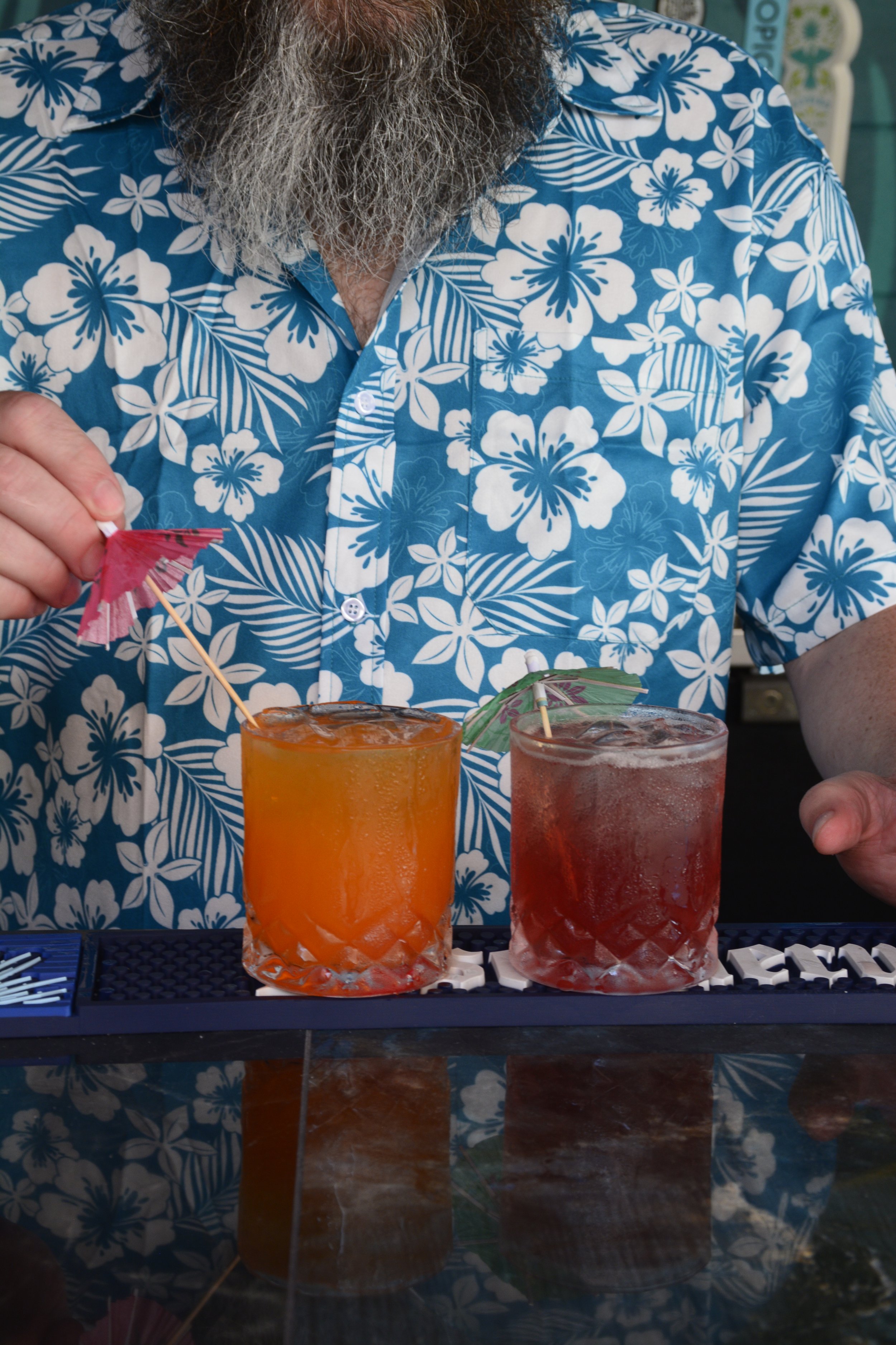 A man with a gray beard wearing a blue Hawaiian shirt with white flowers, holding a pink paper umbrella, standing behind two fruity drinks on a bar counter with a black rubber mat.