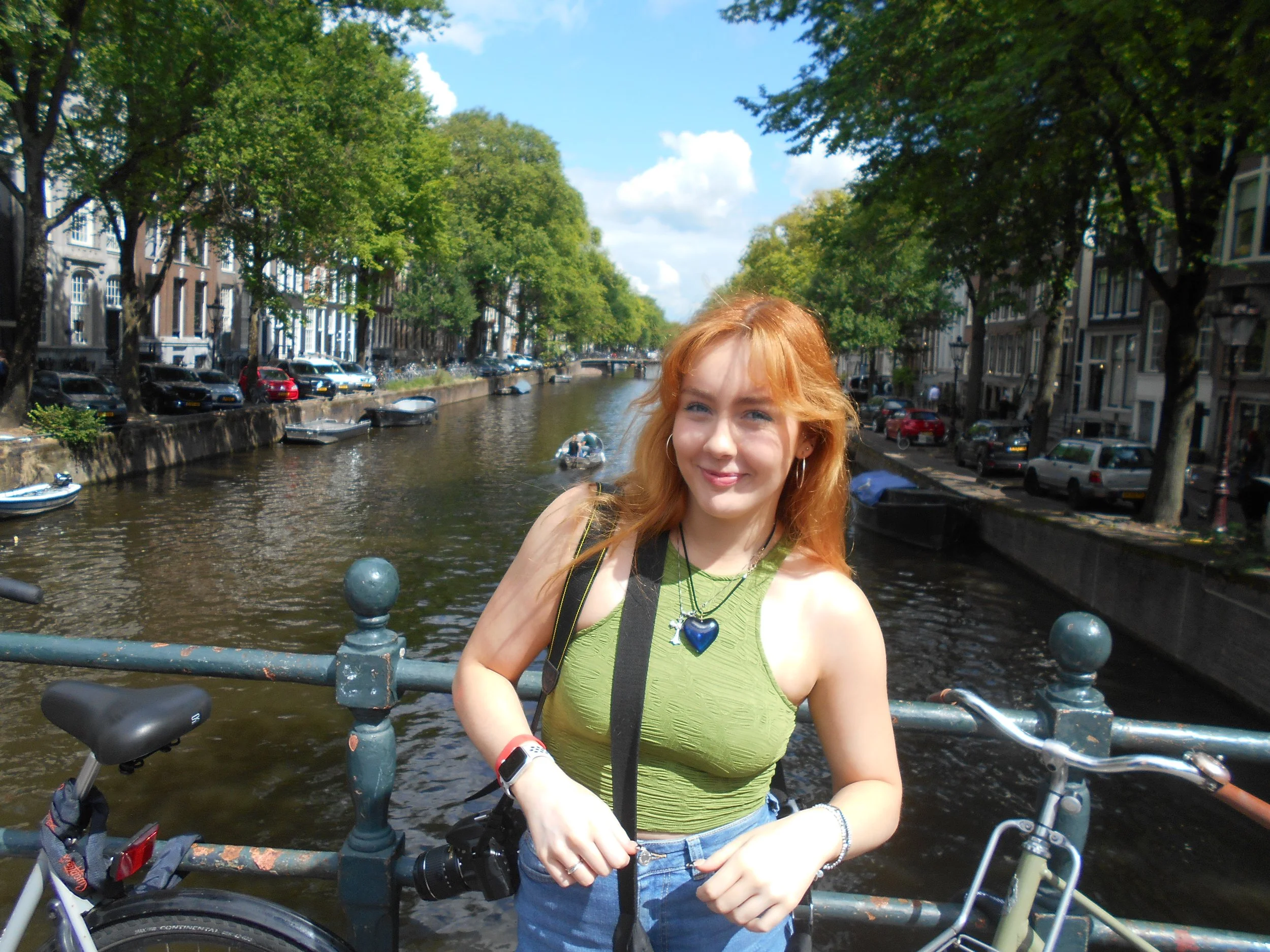 A young woman with red hair smiling in front of a canal with boats and trees, holding the handlebars of a bicycle.