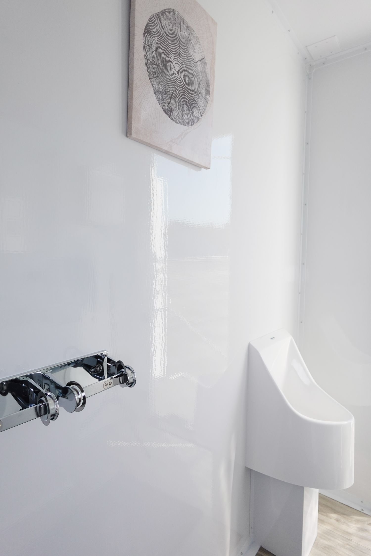 A white bathroom with a white urinal, a wall-mounted paper towel dispenser, and a painting of a tree trunk cross section on the wall.
