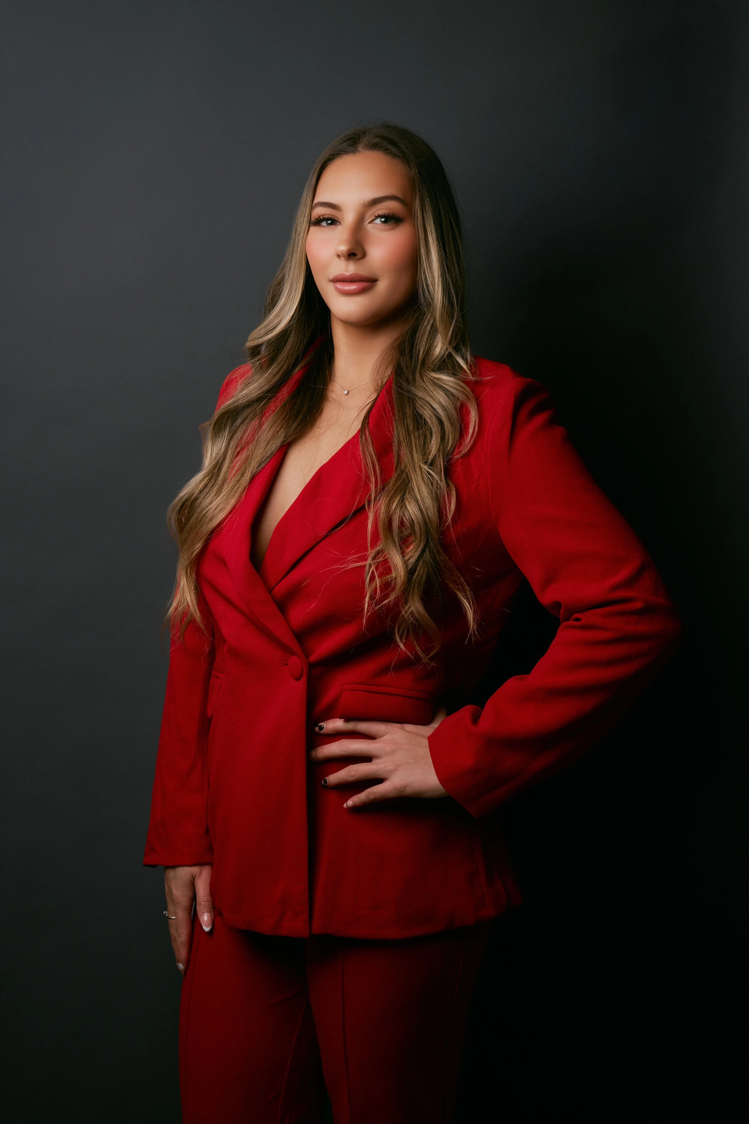 A woman with long wavy hair wearing a red suit and matching pants, standing against a dark background.