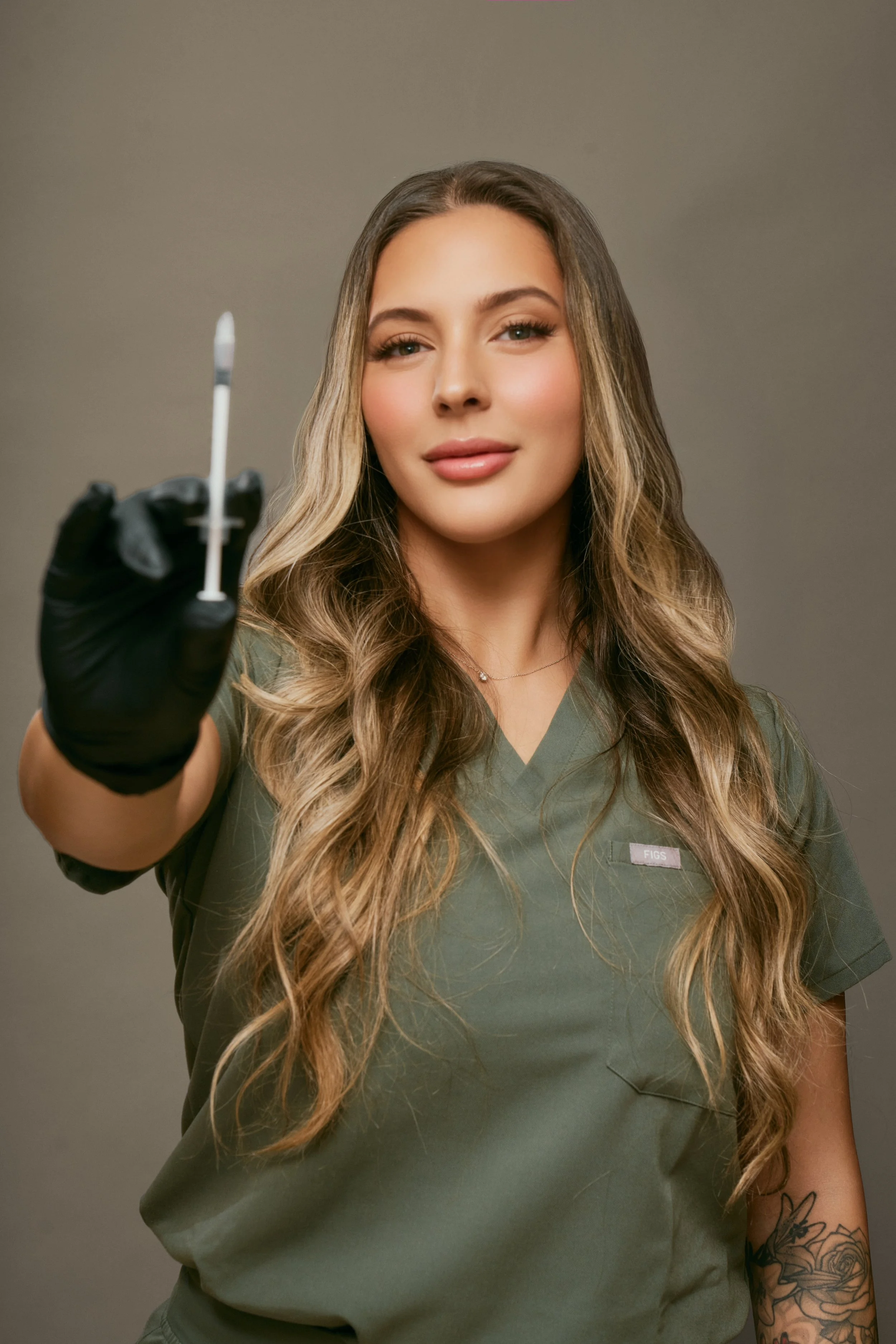 A woman with long wavy hair and tattoos on her arm holds a swab stick in front of her face, with a neutral gray background.