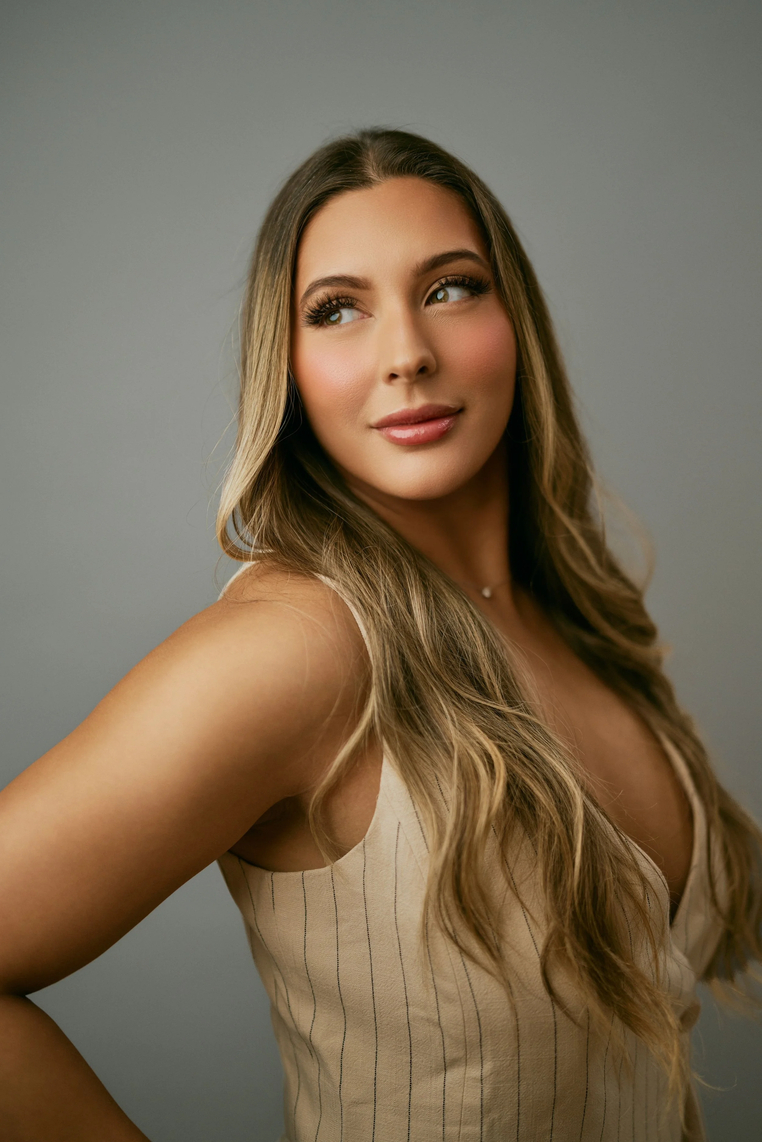 Close-up of a smiling woman with long, wavy hair wearing a beige sleeveless top with thin vertical stripes, posing against a plain gray background.
