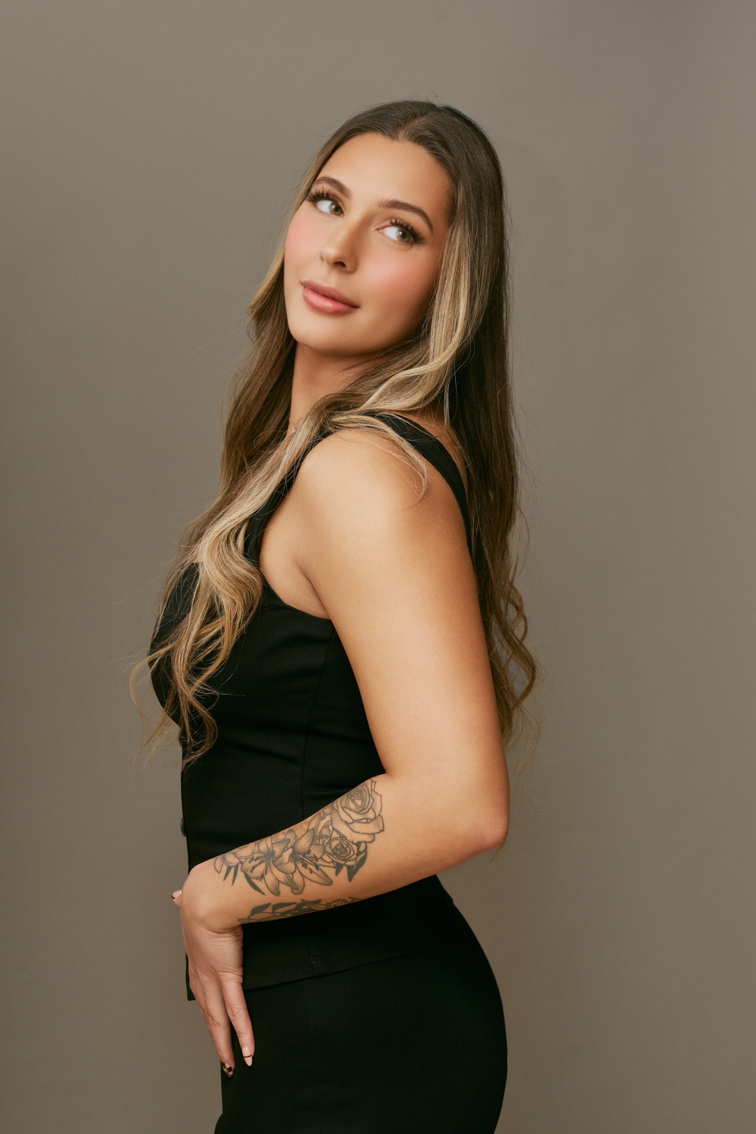 A woman with long, wavy brown and blonde hair, wearing a black sleeveless dress, standing against a plain gray background.